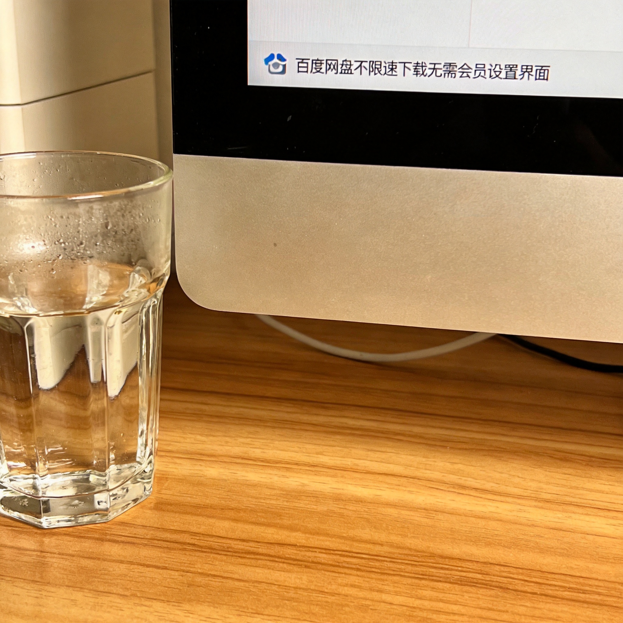 书桌上的主机，屏幕显示百度网盘不限速下载无需会员设置界面，桌角放着水杯，自然光