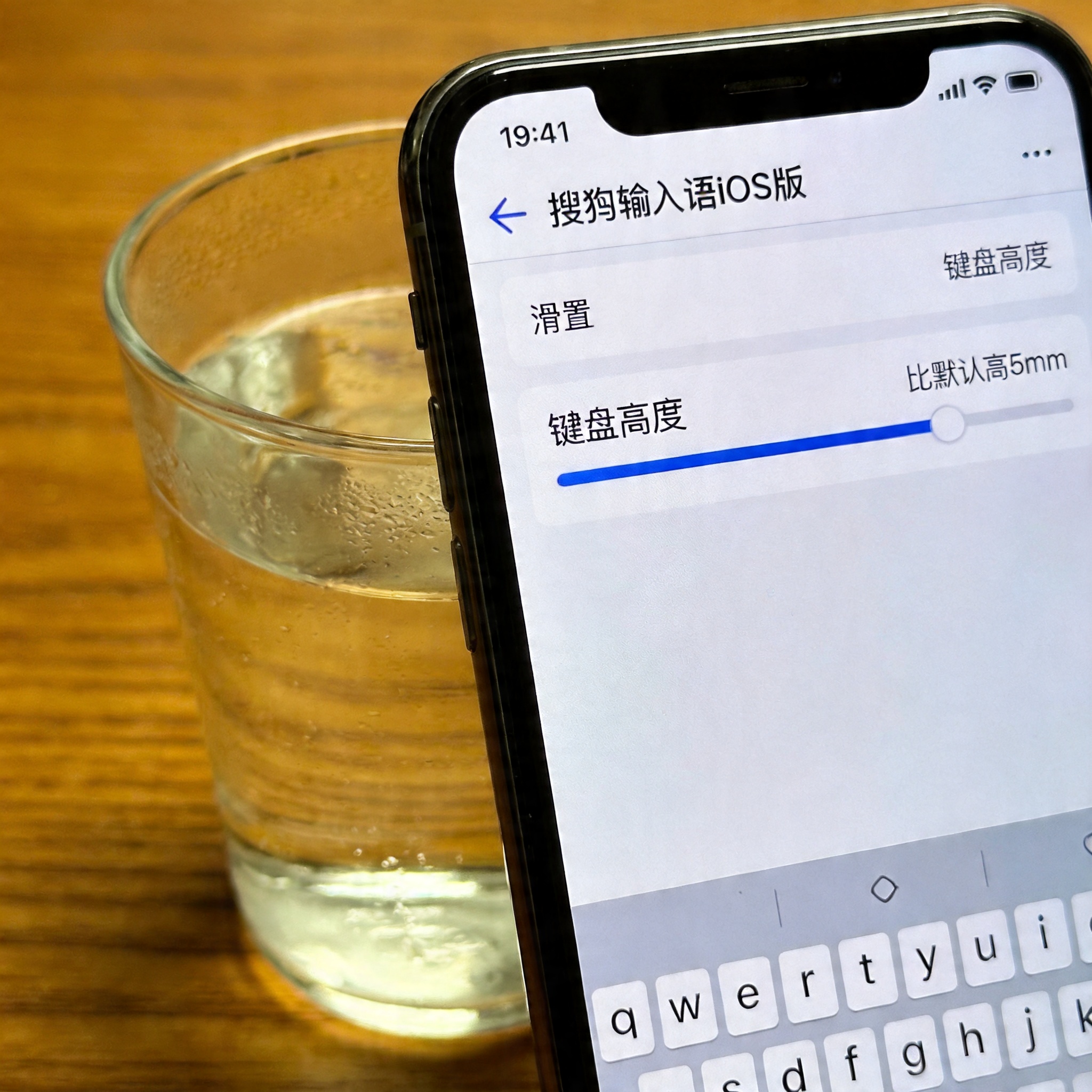 桌面的iPhone屏幕显示搜狗输入法iOS版的键盘设置界面，滑块停在比默认高5mm的位置，旁边放着半杯温水