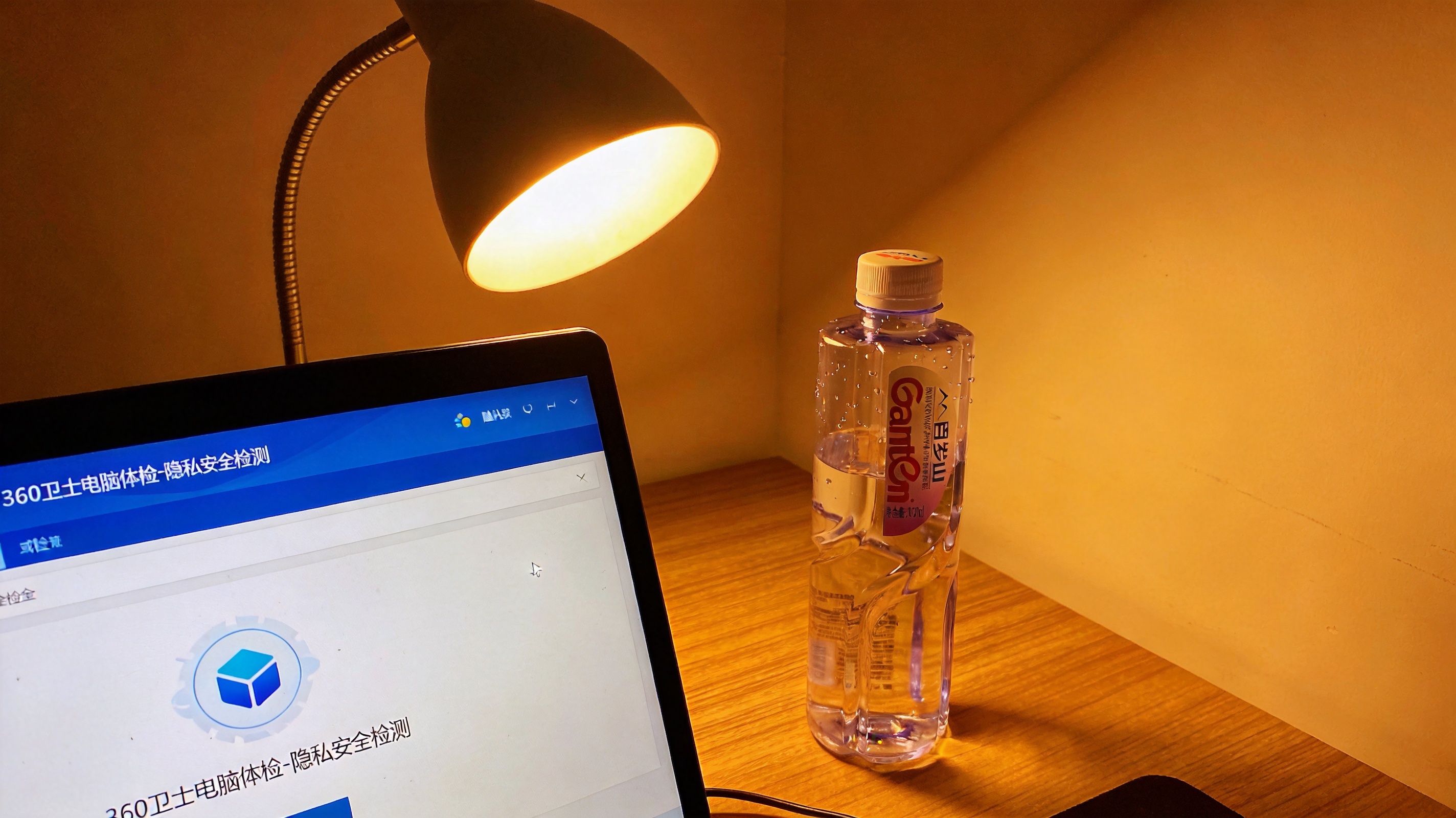 电脑屏幕显示360卫士电脑体检的隐私安全检测页面，桌面有半瓶矿泉水，台灯暖光