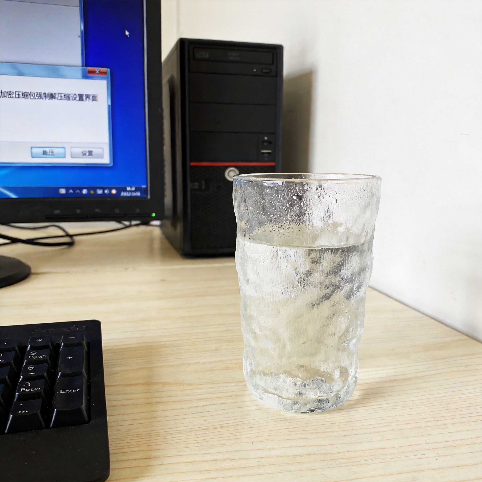 书桌上的主机，屏幕显示加密压缩包强制解压缩设置界面，桌角放着水杯，自然光