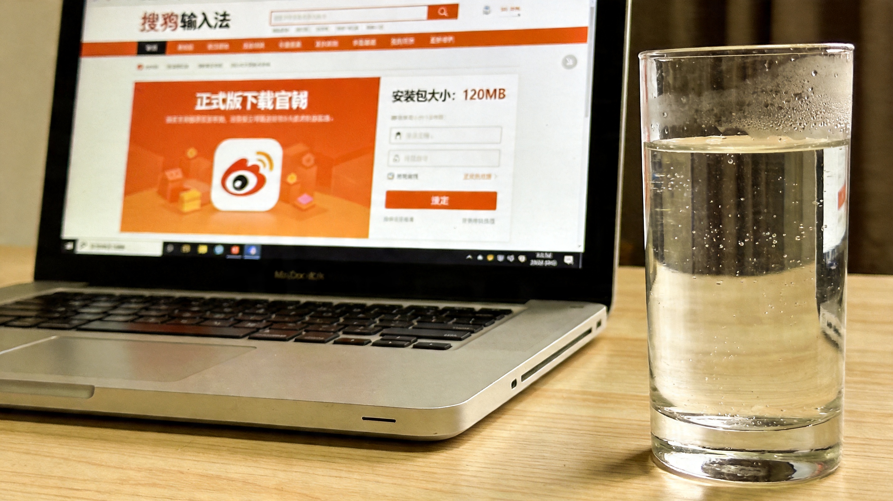 电脑桌面，屏幕显示搜狗输入法官网的正式版下载页面，页面标注安装包大小120MB，旁边放着半杯凉白开，室内自然光