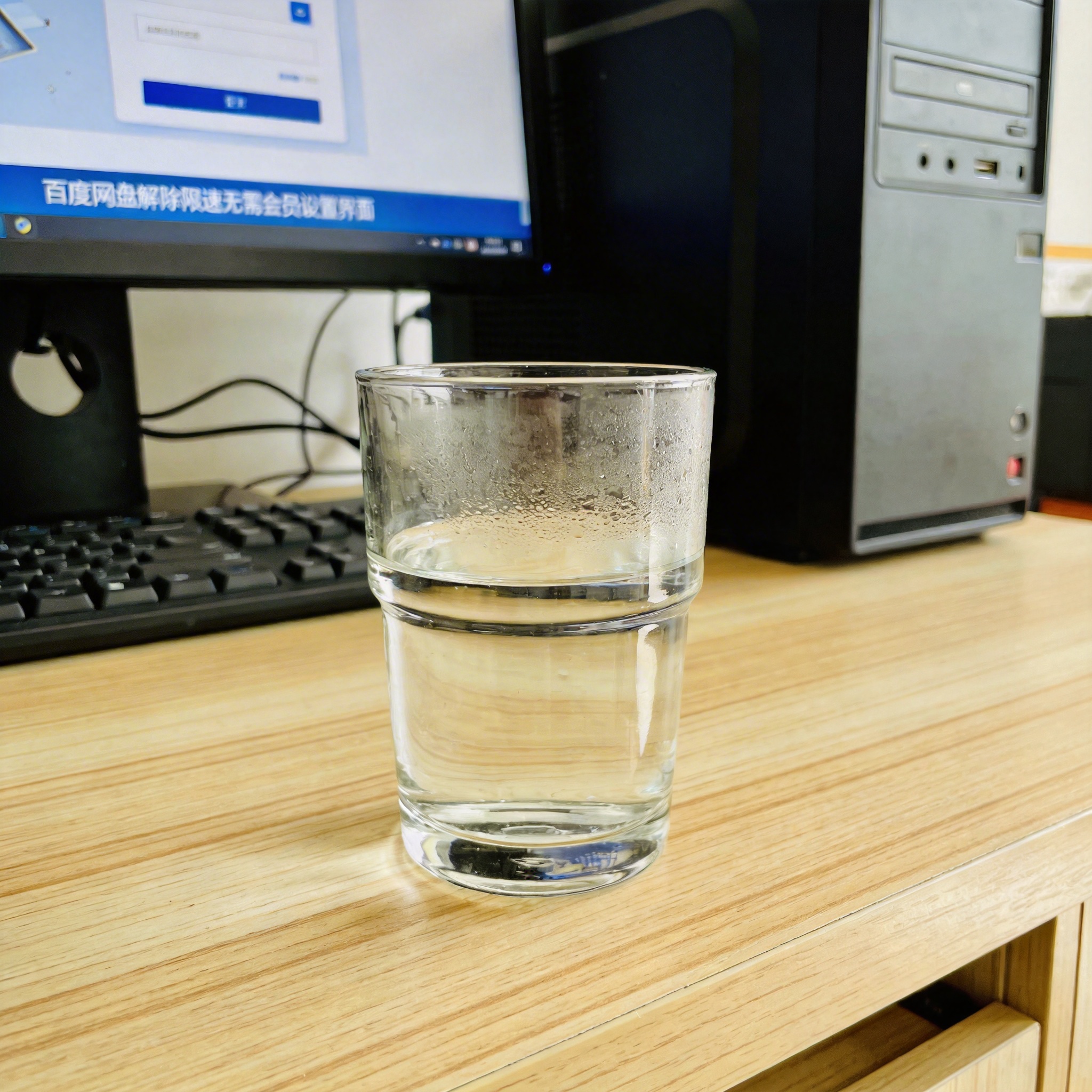 书桌上的主机，屏幕显示百度网盘解除限速无需会员设置界面，桌角放着水杯，自然光