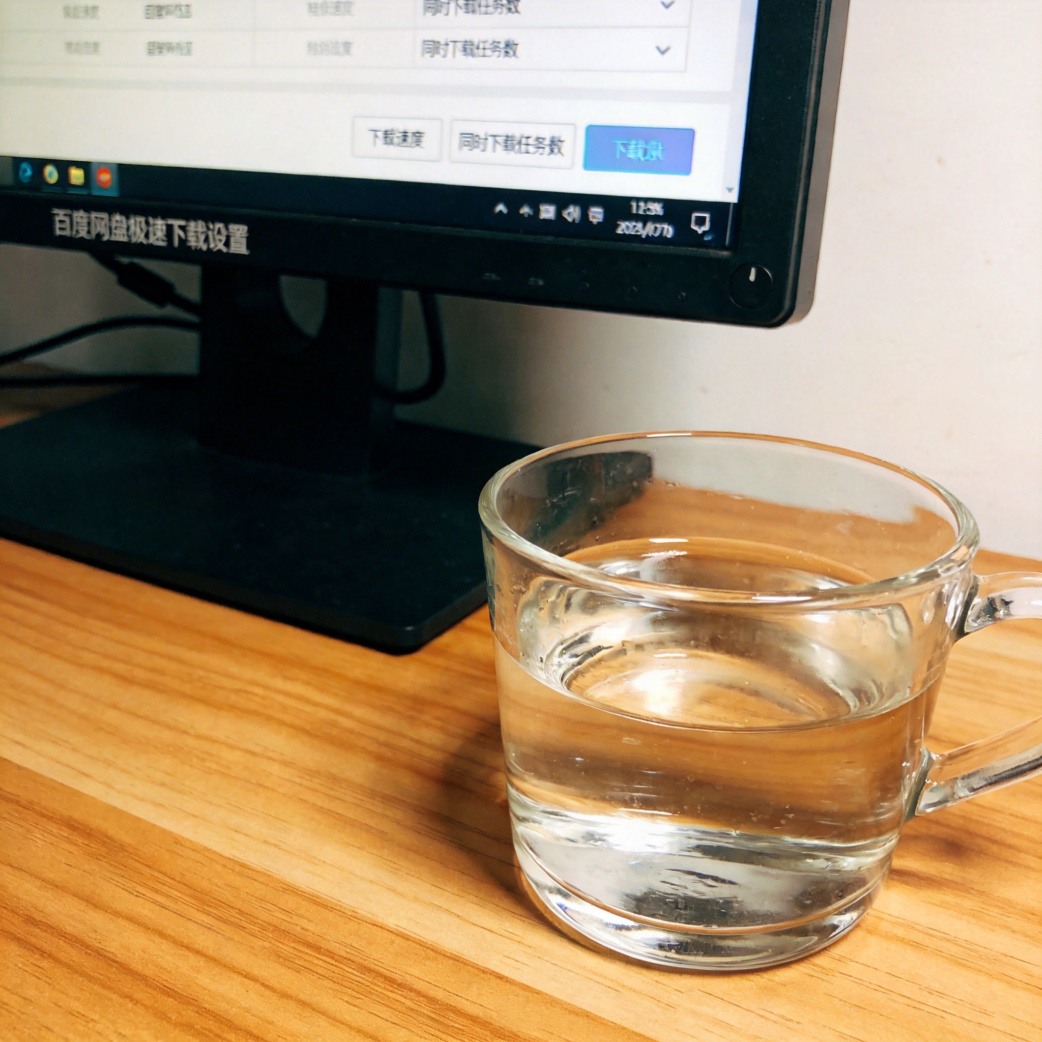 书桌上的主机，屏幕显示百度网盘极速下载设置设置界面，桌角放着水杯，自然光