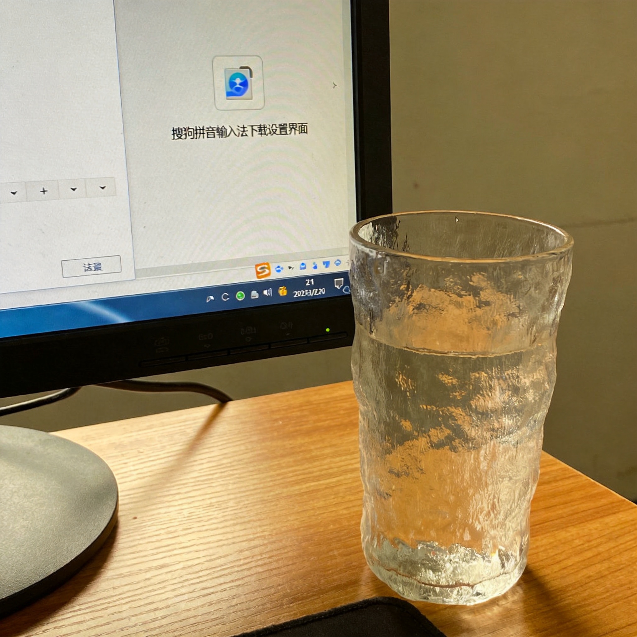 书桌上的主机，屏幕显示搜狗拼音输入法下载设置界面，桌角放着水杯，自然光