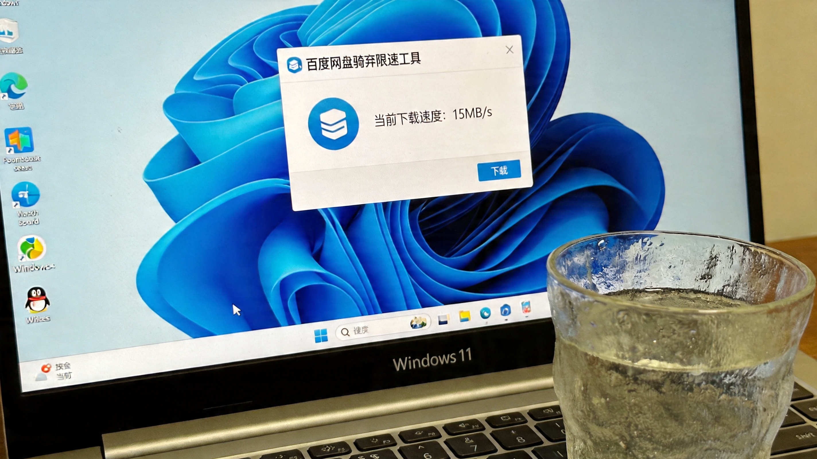 Windows11系统的电脑桌面，屏幕显示百度网盘破解限速工具的下载界面，当前下载速度15MB/s，桌面角落放着半杯温水，室内自然光