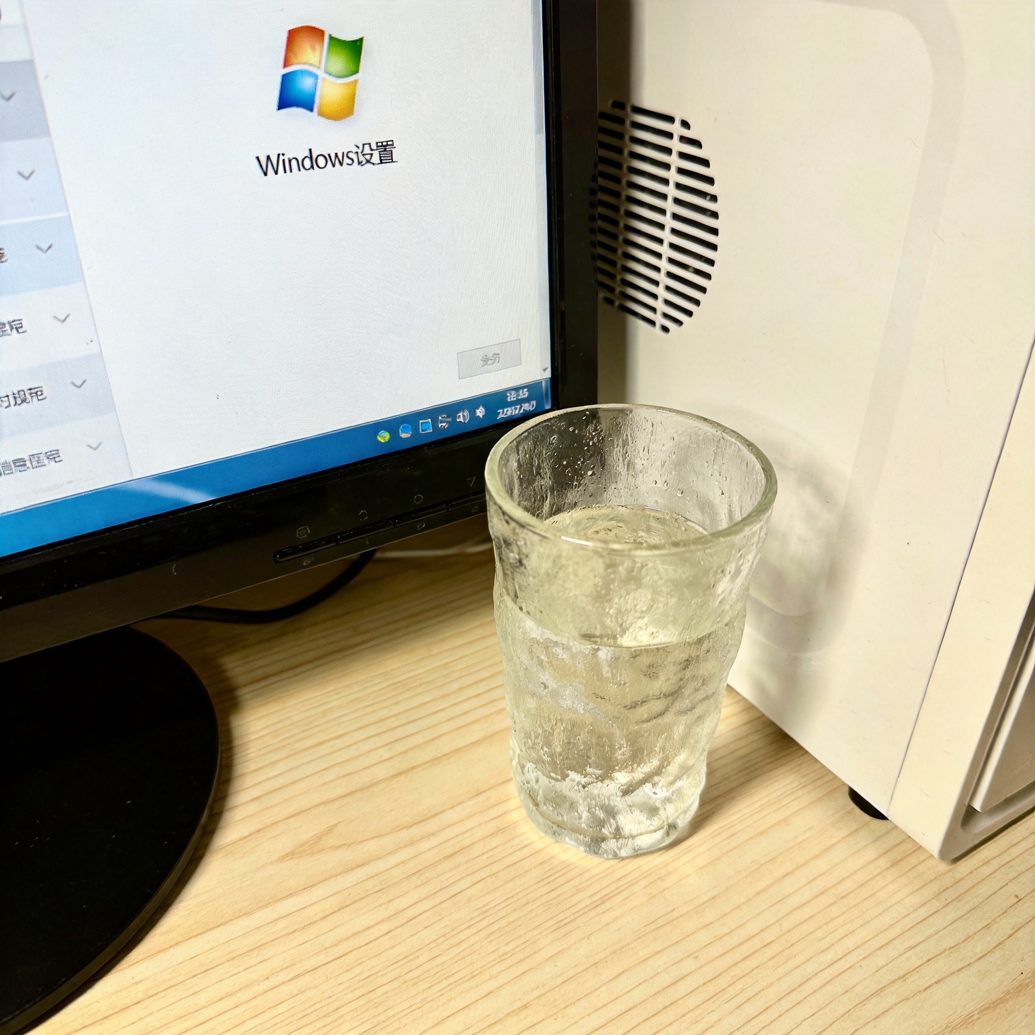 书桌上的主机，屏幕显示Windows设置界面，桌角放着水杯，自然光