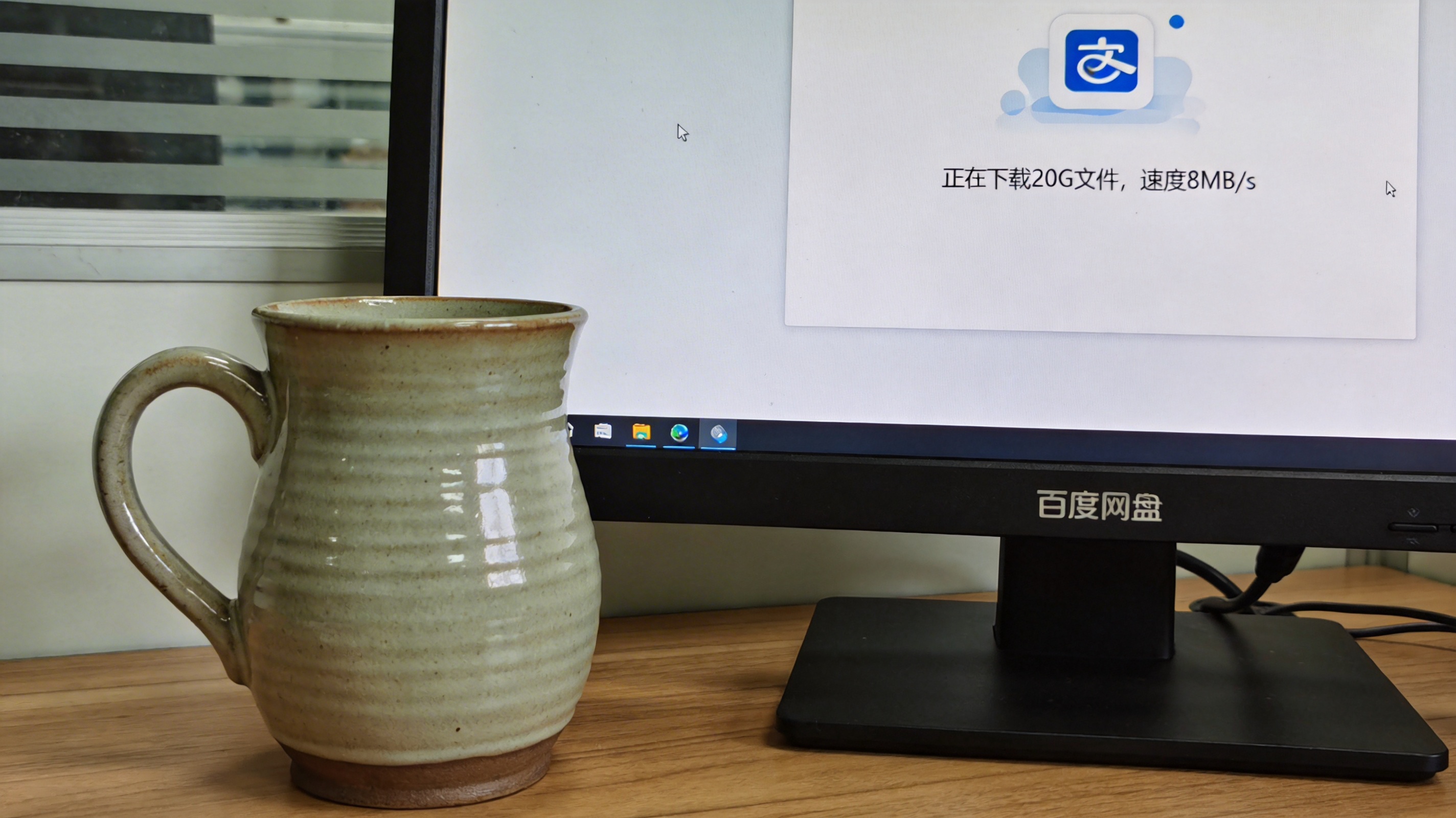 电脑桌面的显示器，屏幕显示百度网盘界面，正在下载20G大小的文件，当前速度显示8MB/s，旁边放着陶瓷水杯，室内自然光