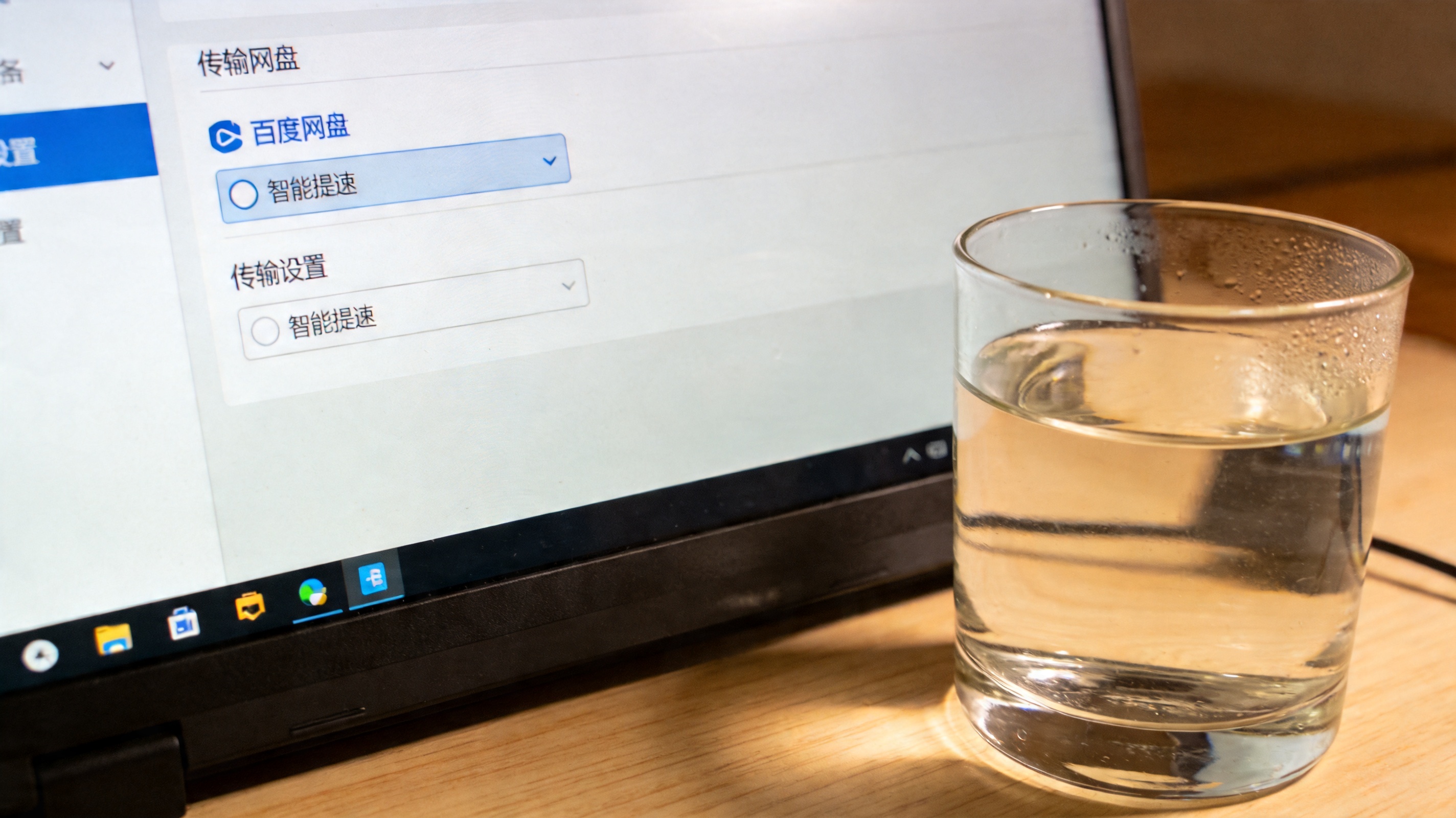 电脑桌面，屏幕显示百度网盘客户端传输设置界面，“智能提速”选项已勾选，旁边放着半杯温水，室内自然光
