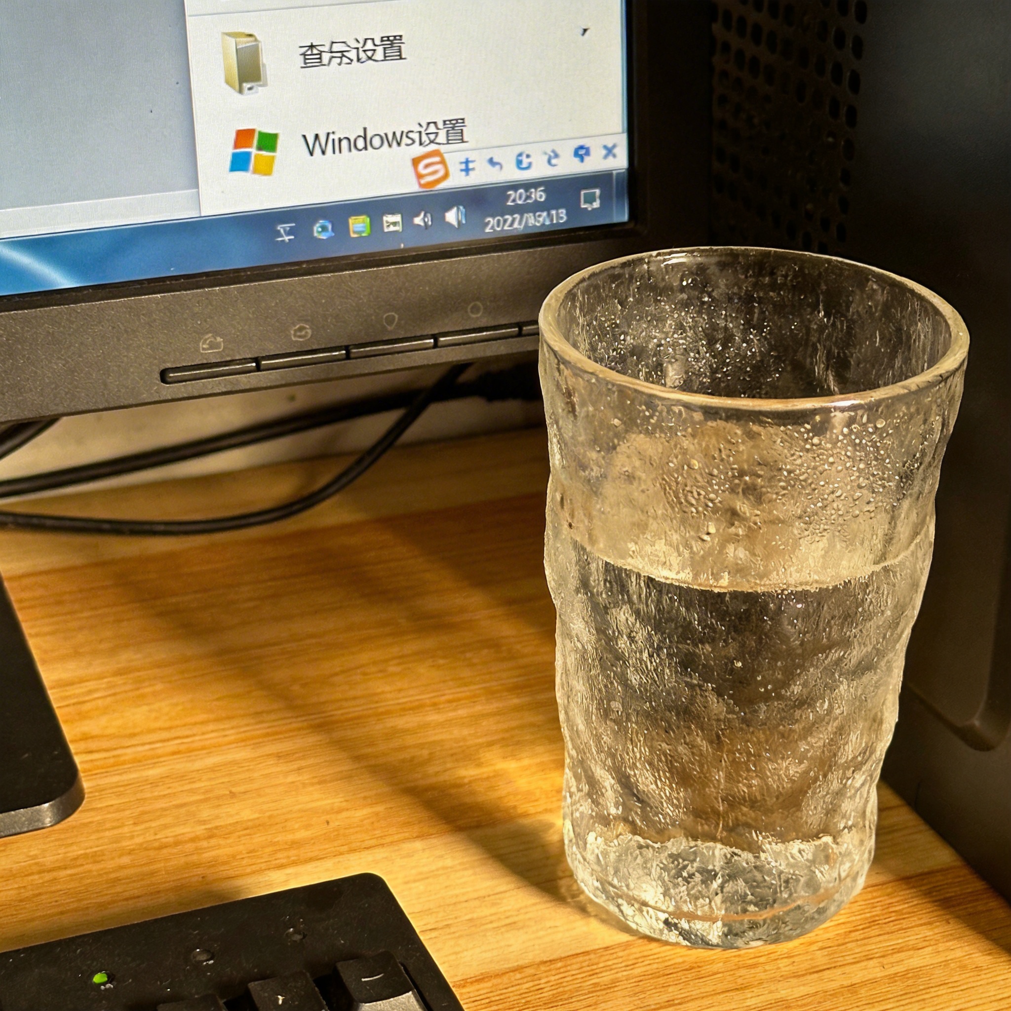 书桌上的主机，屏幕显示Windows设置界面，桌角放着水杯，自然光