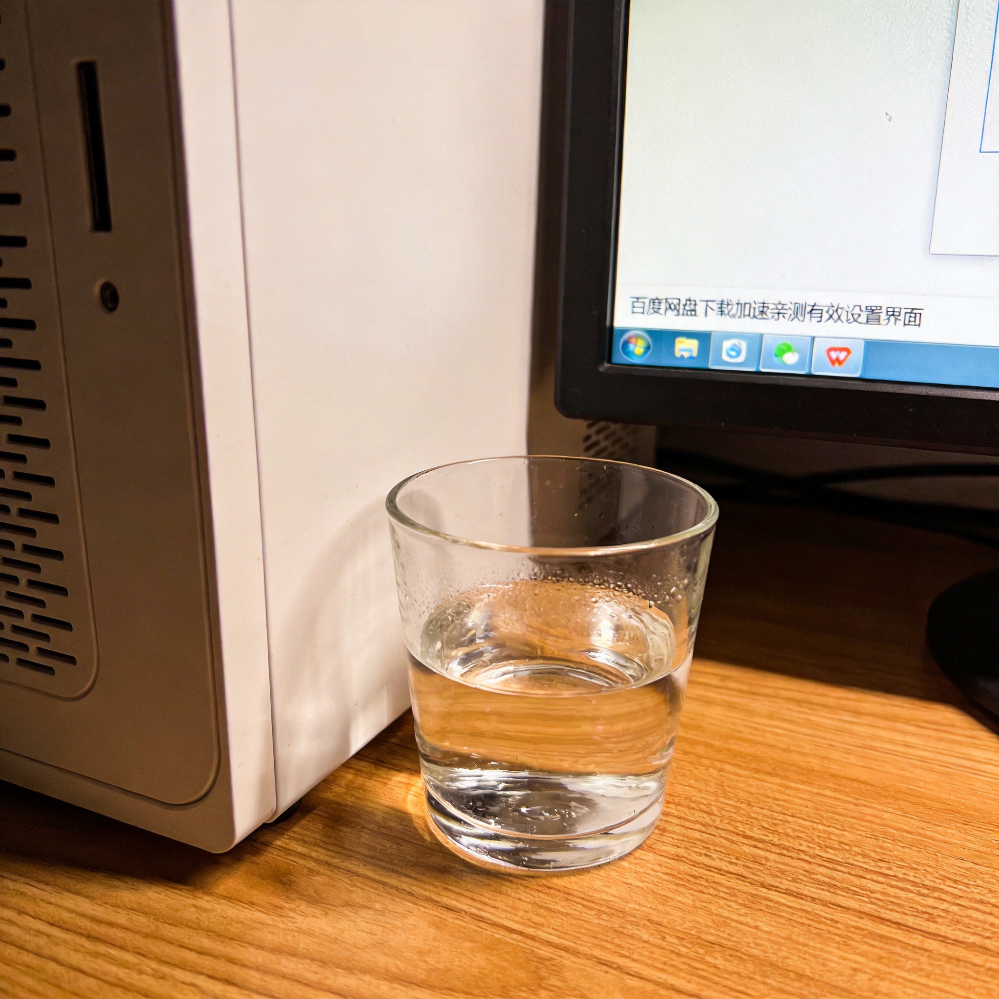 书桌上的主机，屏幕显示百度网盘下载加速亲测有效设置界面，桌角放着水杯，自然光