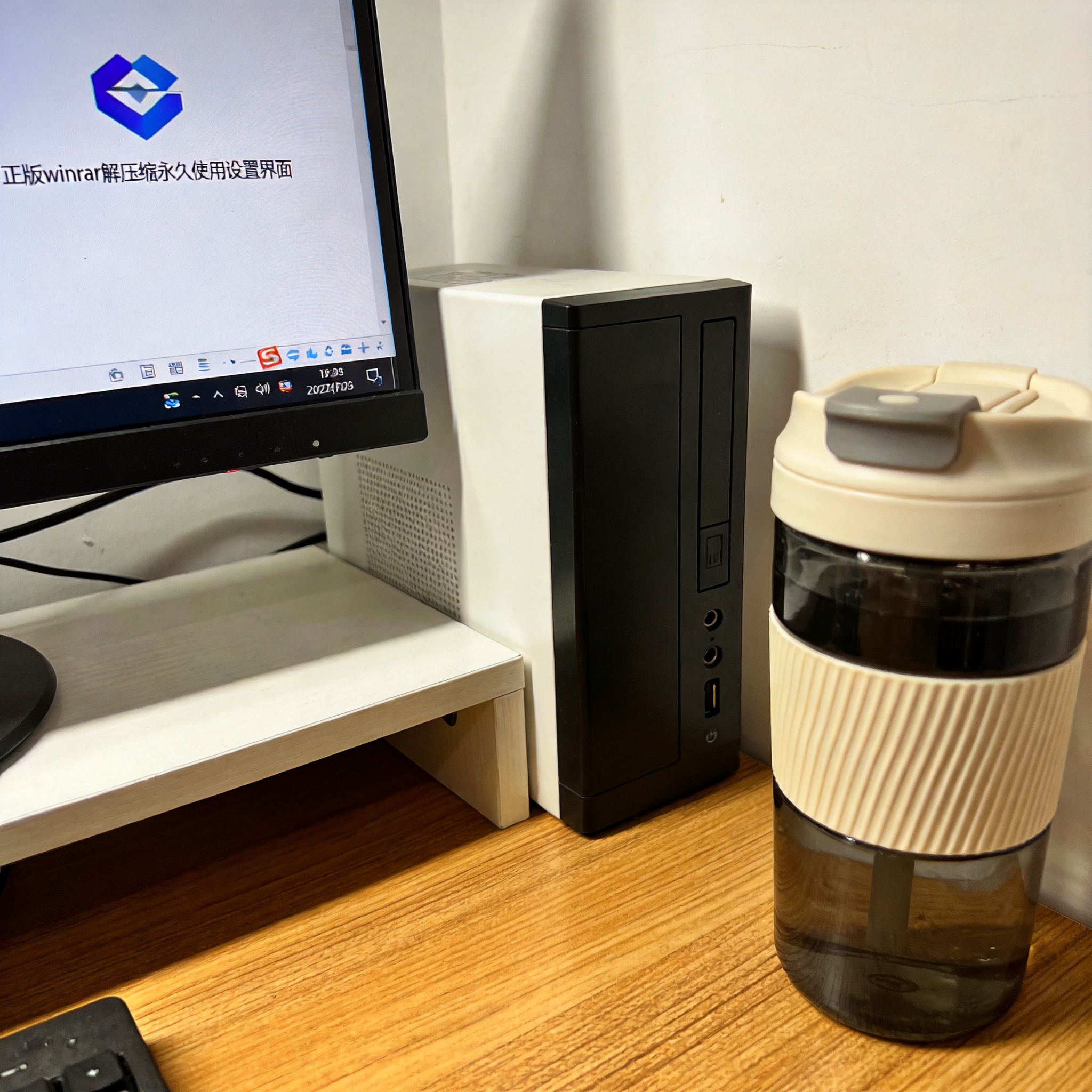 书桌上的主机，屏幕显示正版 winrar 解压缩永久使用设置界面，桌角放着水杯，自然光