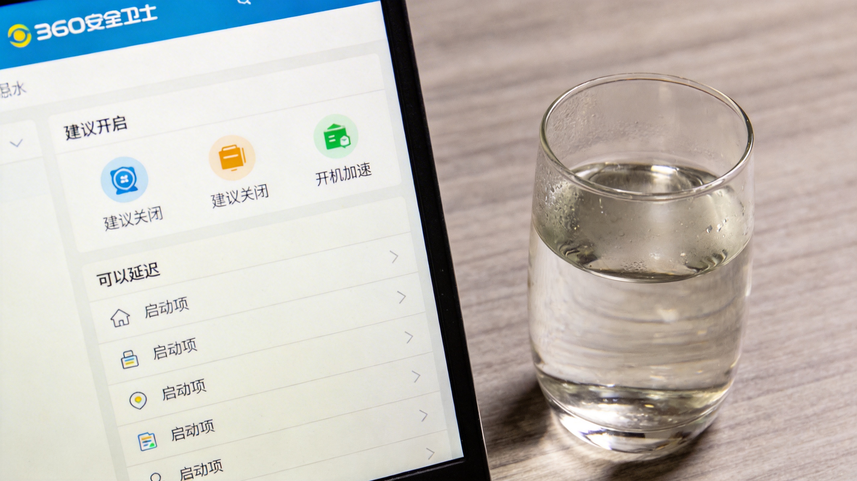电脑桌面，屏幕显示360安全卫士开机加速界面，界面上列着“建议开启”“建议关闭”“可以延迟”的启动项分类，桌面放着半杯温水