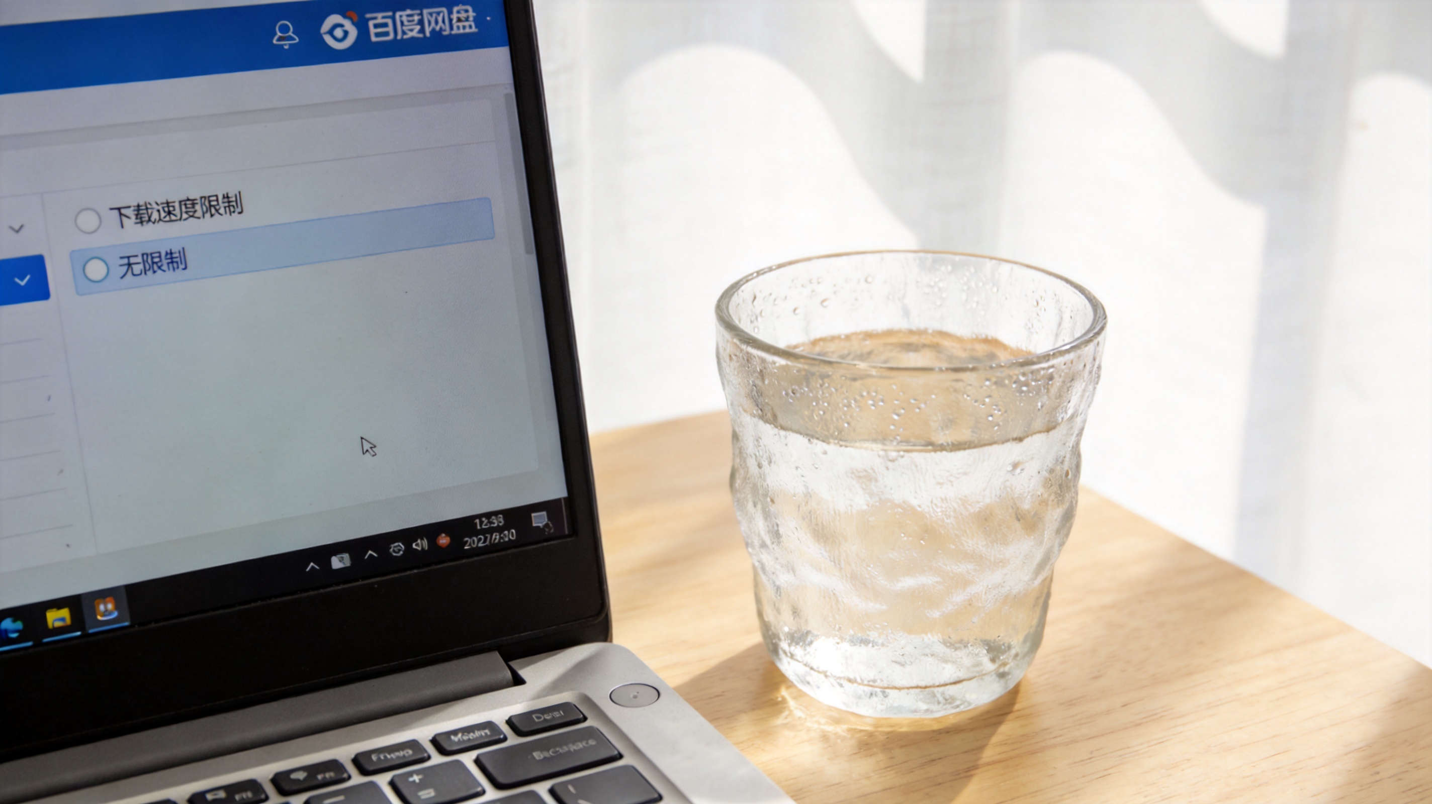 电脑桌面，屏幕显示百度网盘客户端的传输设置页面，“下载速度限制”选项设为“无限制”，旁边放着半杯温水，室内自然光