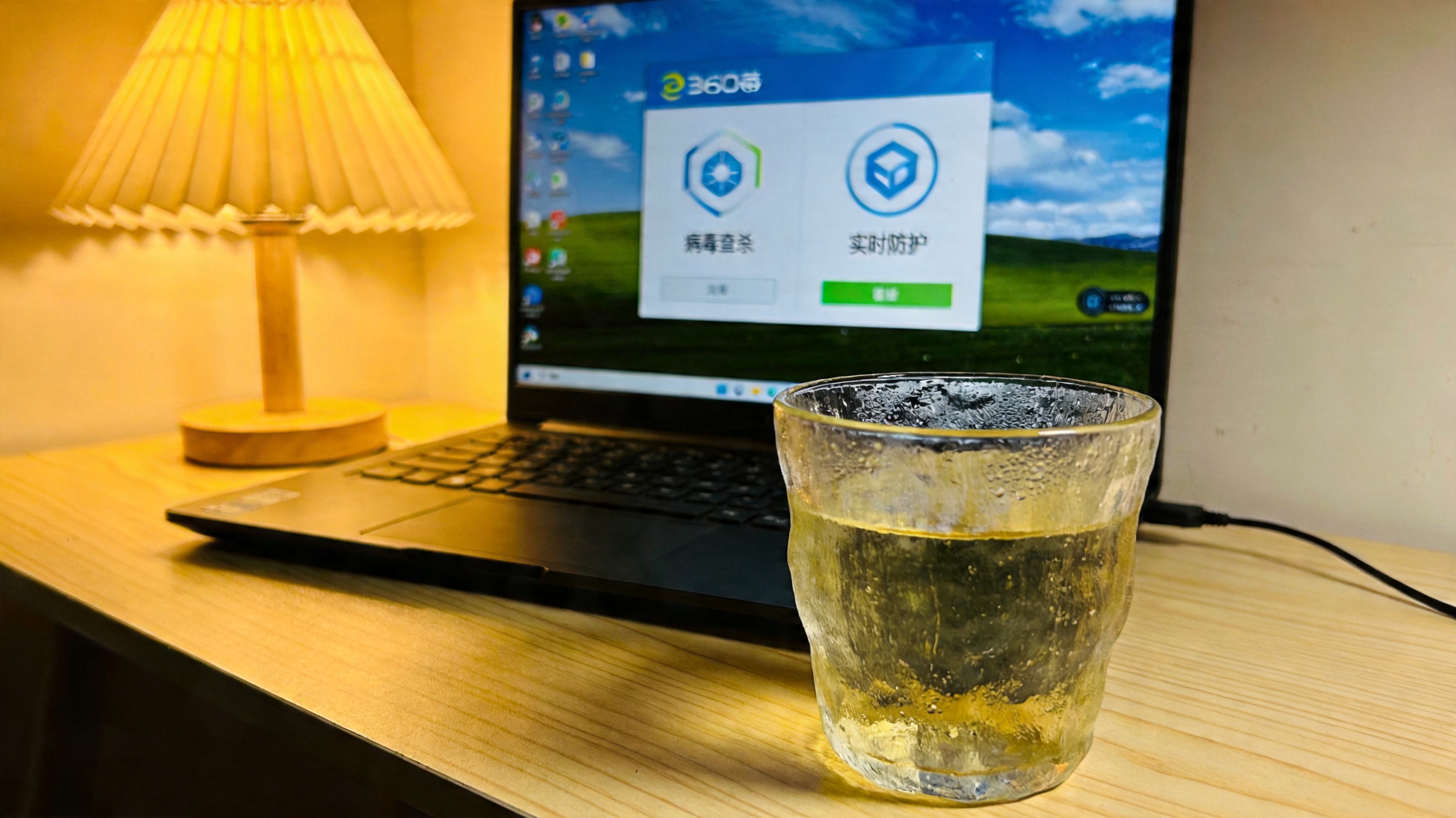 电脑屏幕显示360杀毒界面，界面上有病毒查杀、实时防护选项，旁边的桌面放着半杯温水，台灯暖光