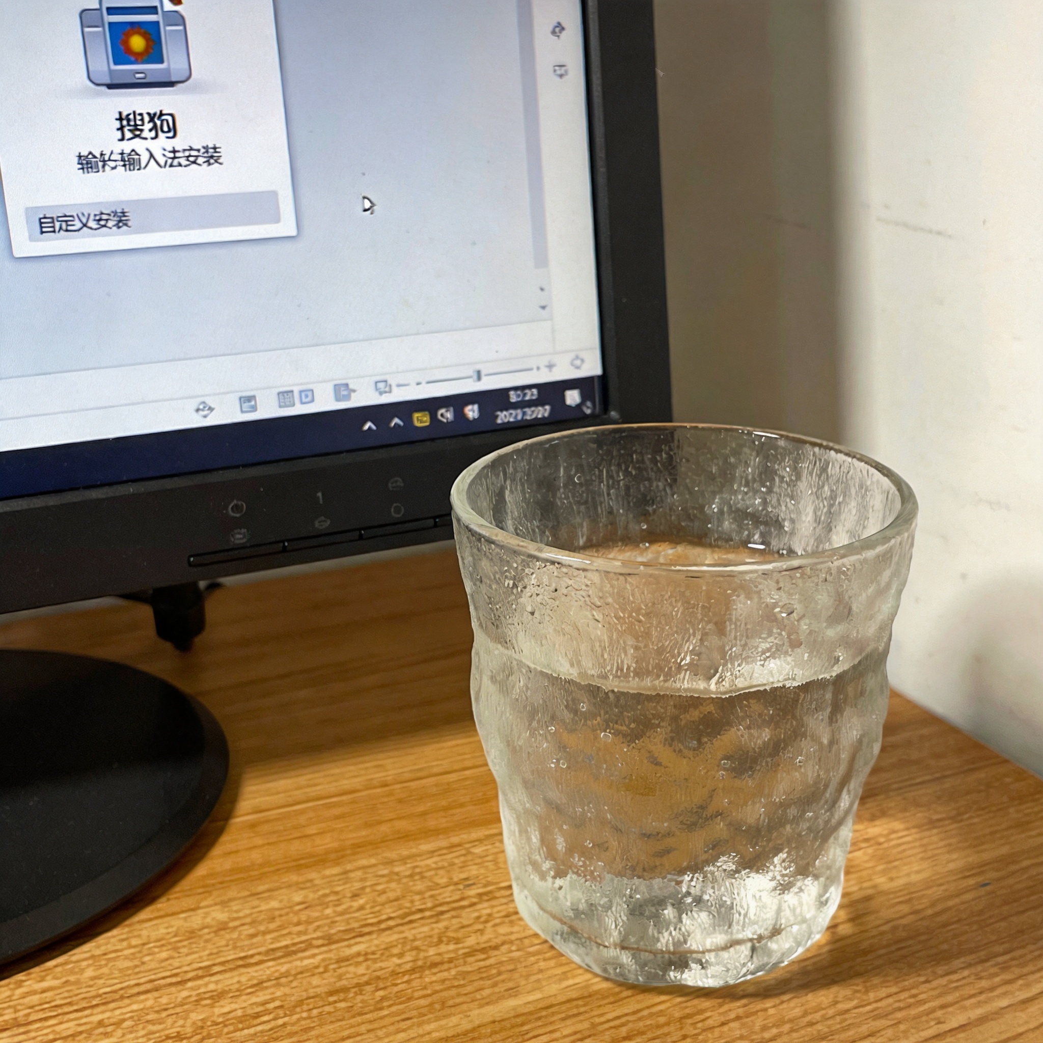 电脑桌的电脑屏幕显示搜狗输入法安装界面，左下角有“自定义安装”选项，旁边放着半杯凉水，室内自然光