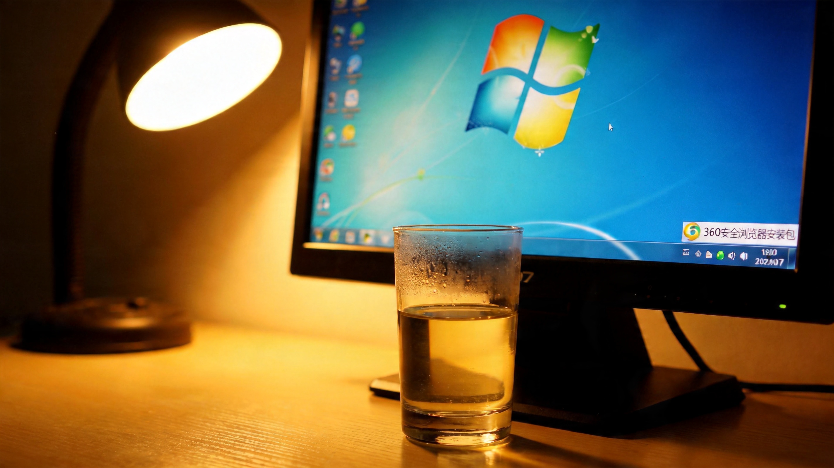 电脑屏幕显示Windows7系统桌面，360安全浏览器安装包放在桌面角落，旁边放着半杯温水，台灯暖光