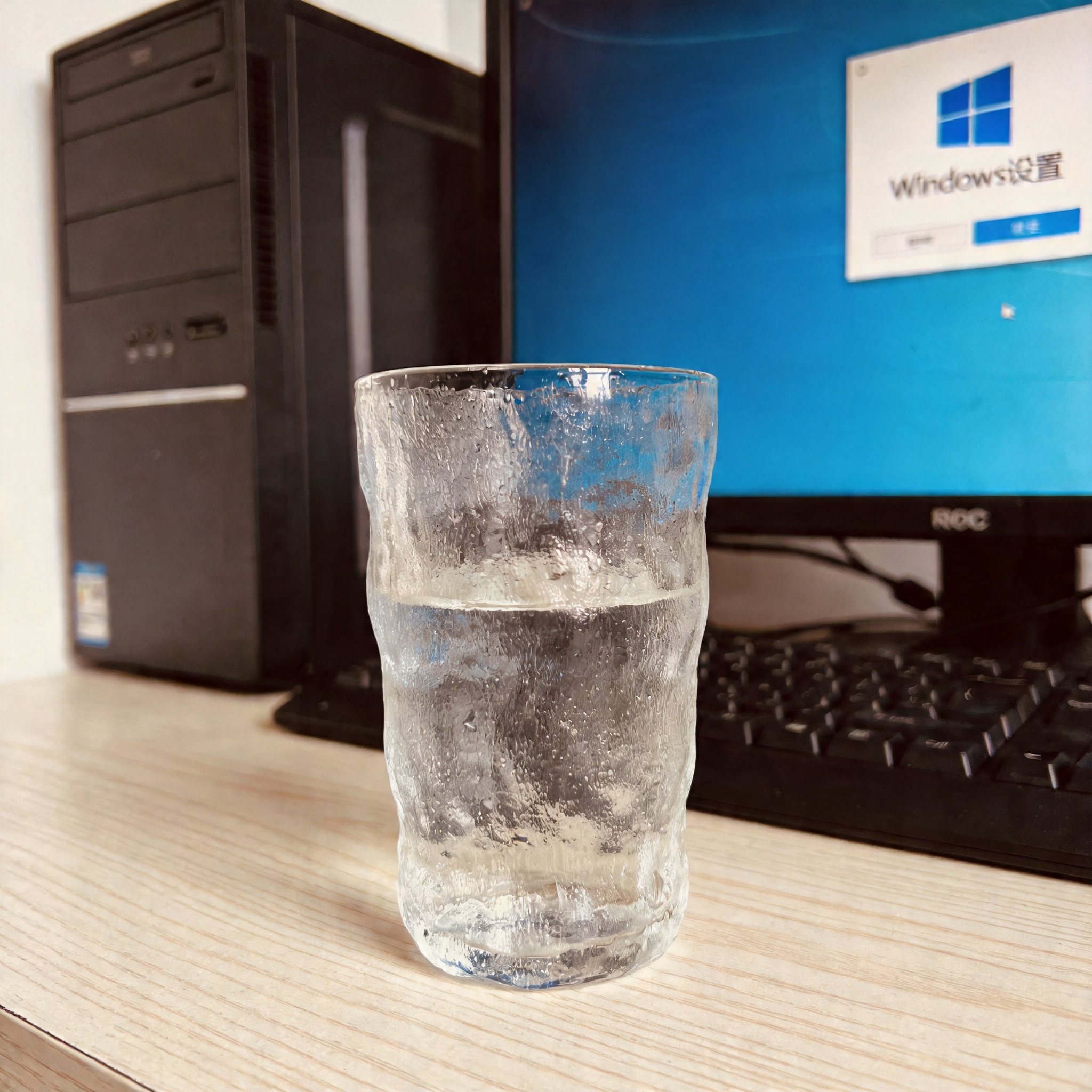 书桌上的主机，屏幕显示Windows设置界面，桌角放着水杯，自然光