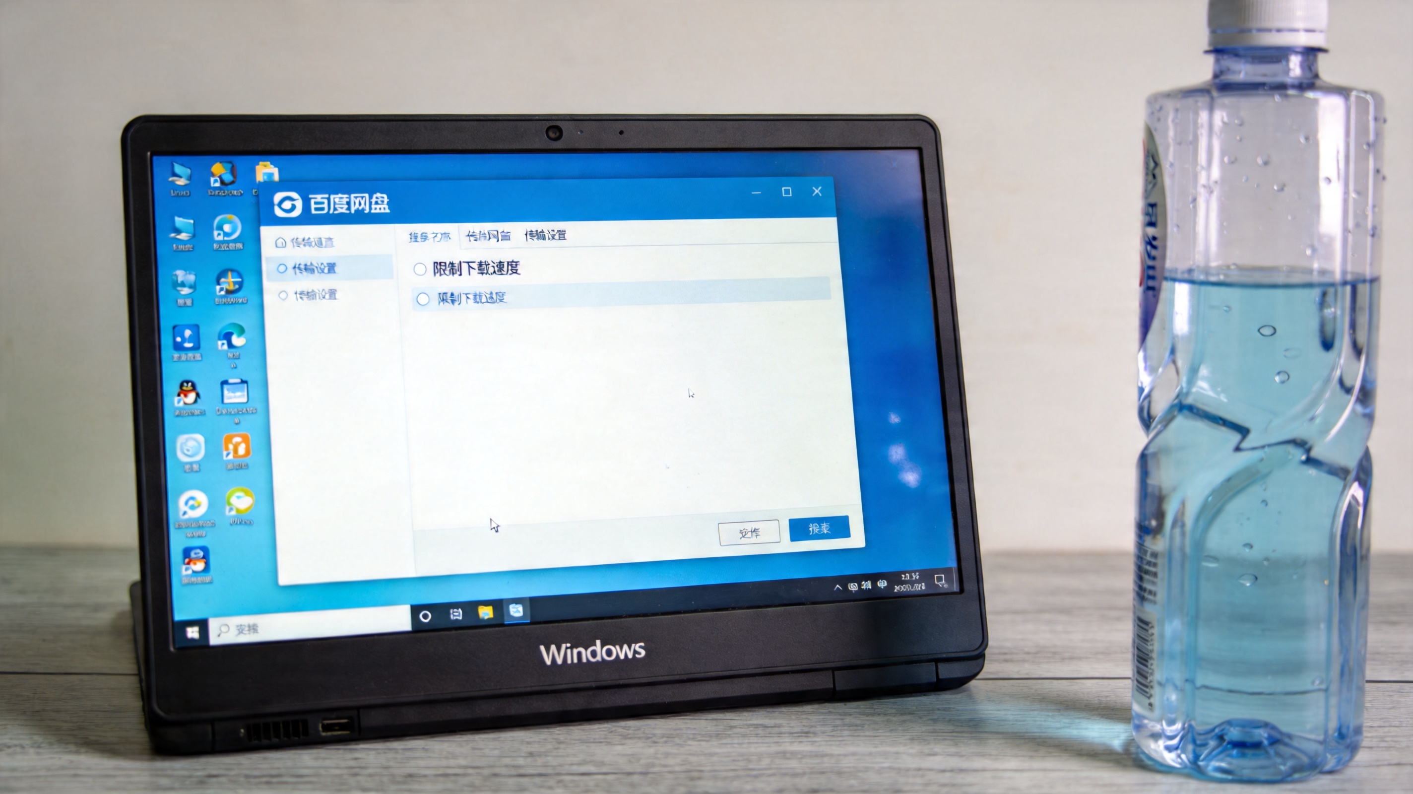 Windows系统的电脑放在桌面上，屏幕显示百度网盘传输设置界面，取消勾选“限制下载速度”选项，桌面放着半瓶矿泉水