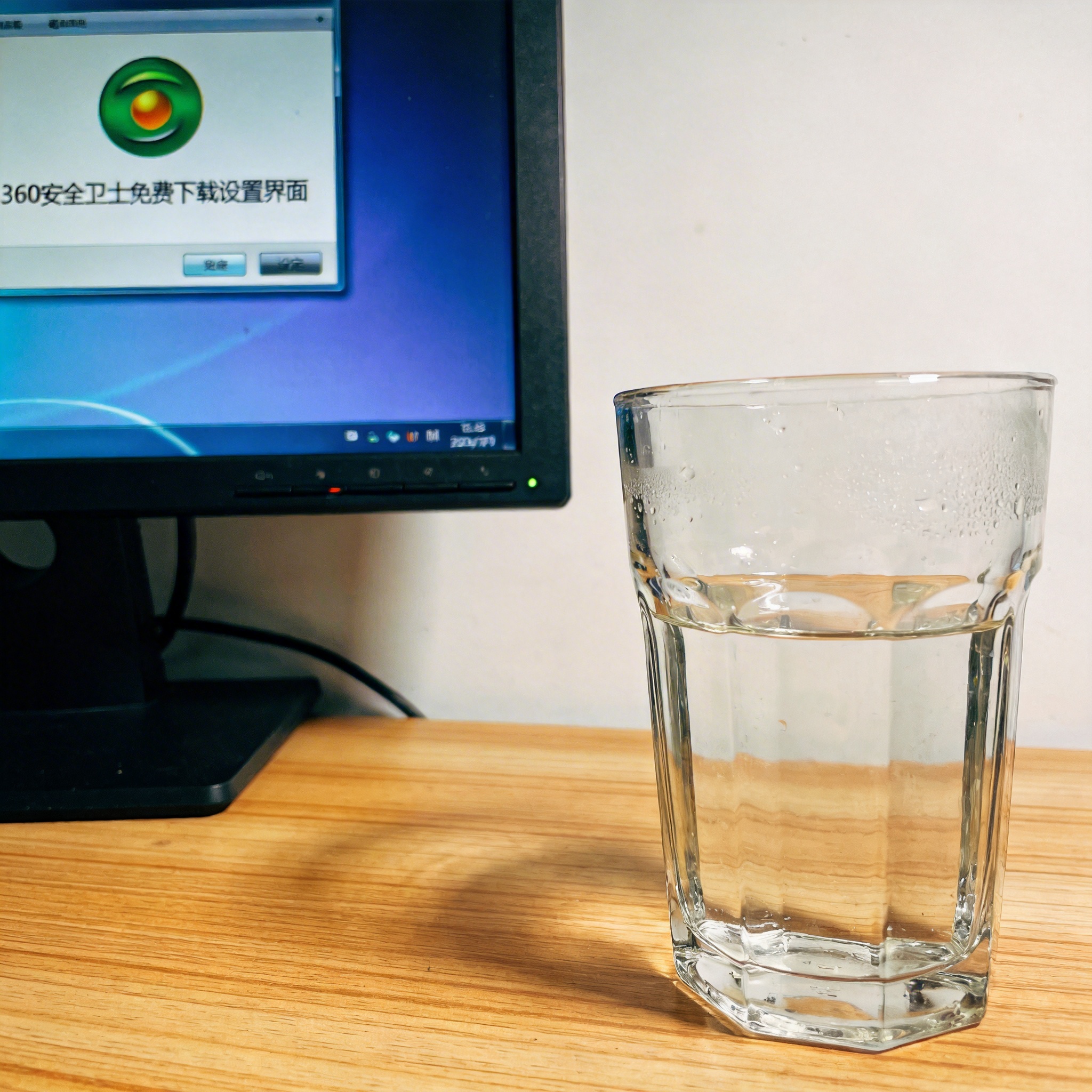 书桌上的主机，屏幕显示360 安全卫士免费下载设置界面，桌角放着水杯，自然光