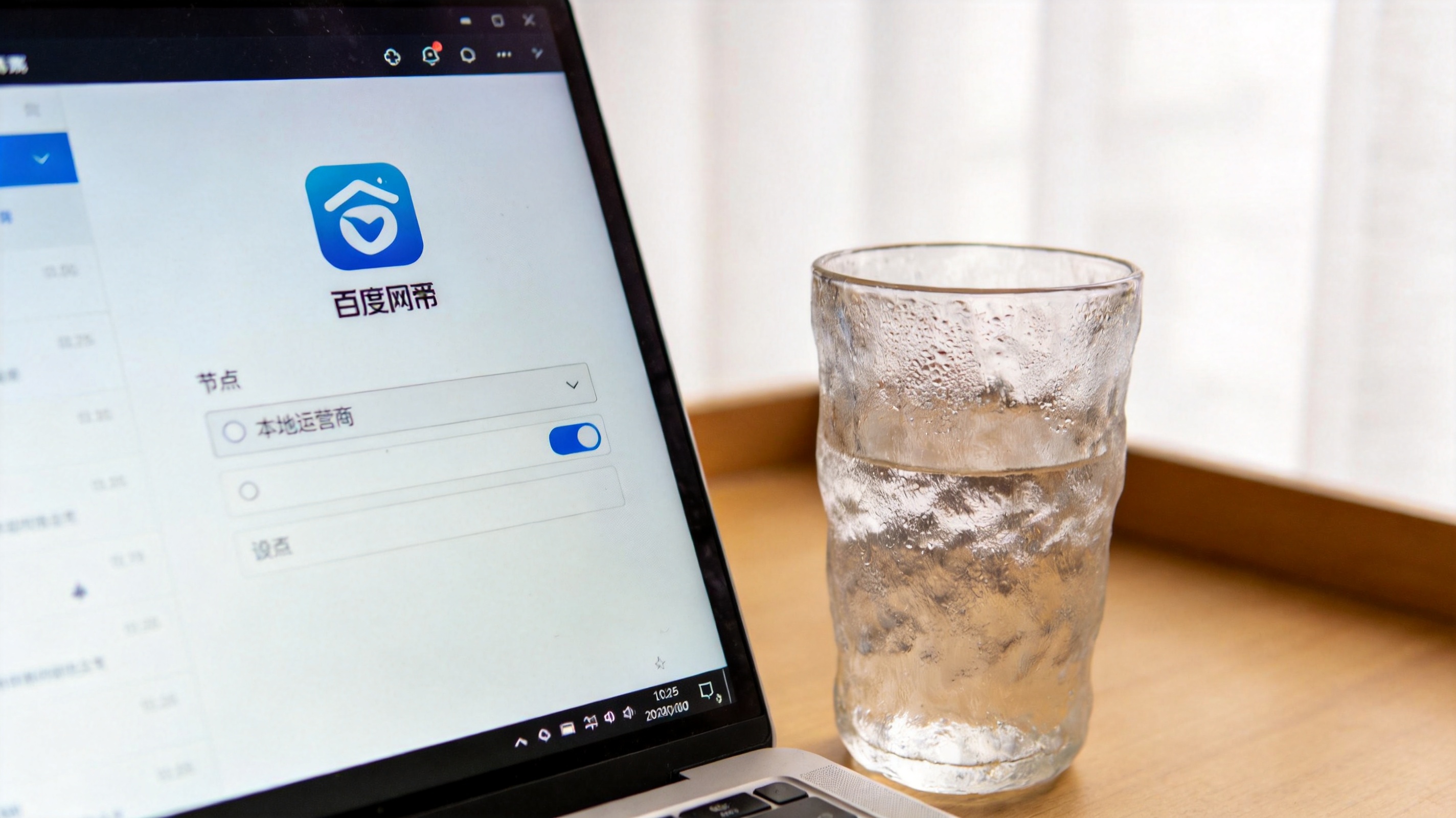 电脑桌面，屏幕显示百度网盘APP的下载设置界面，节点选择栏里显示本地运营商选项，旁边放着半杯温水，室内自然光