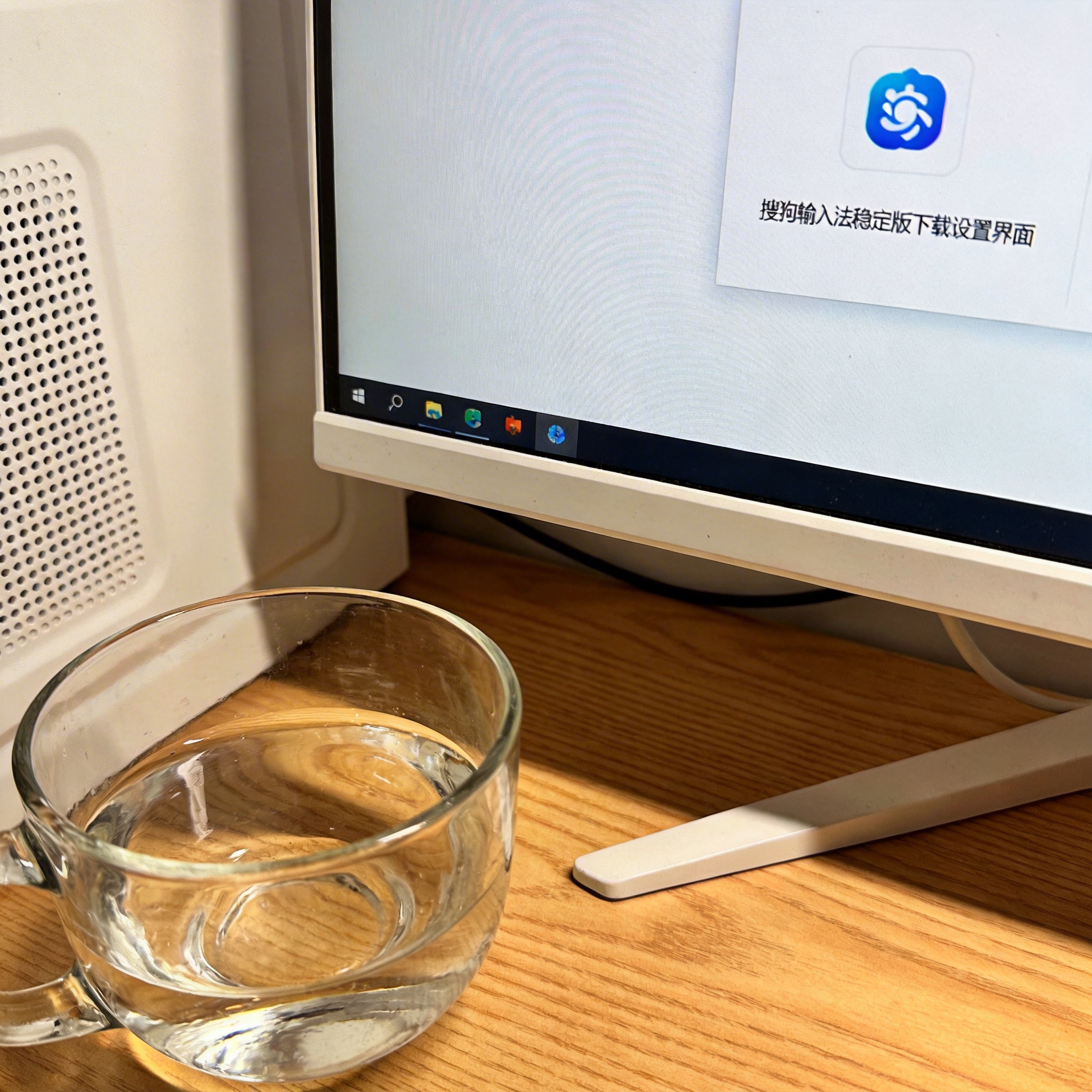 书桌上的主机，屏幕显示搜狗输入法稳定版下载设置界面，桌角放着水杯，自然光
