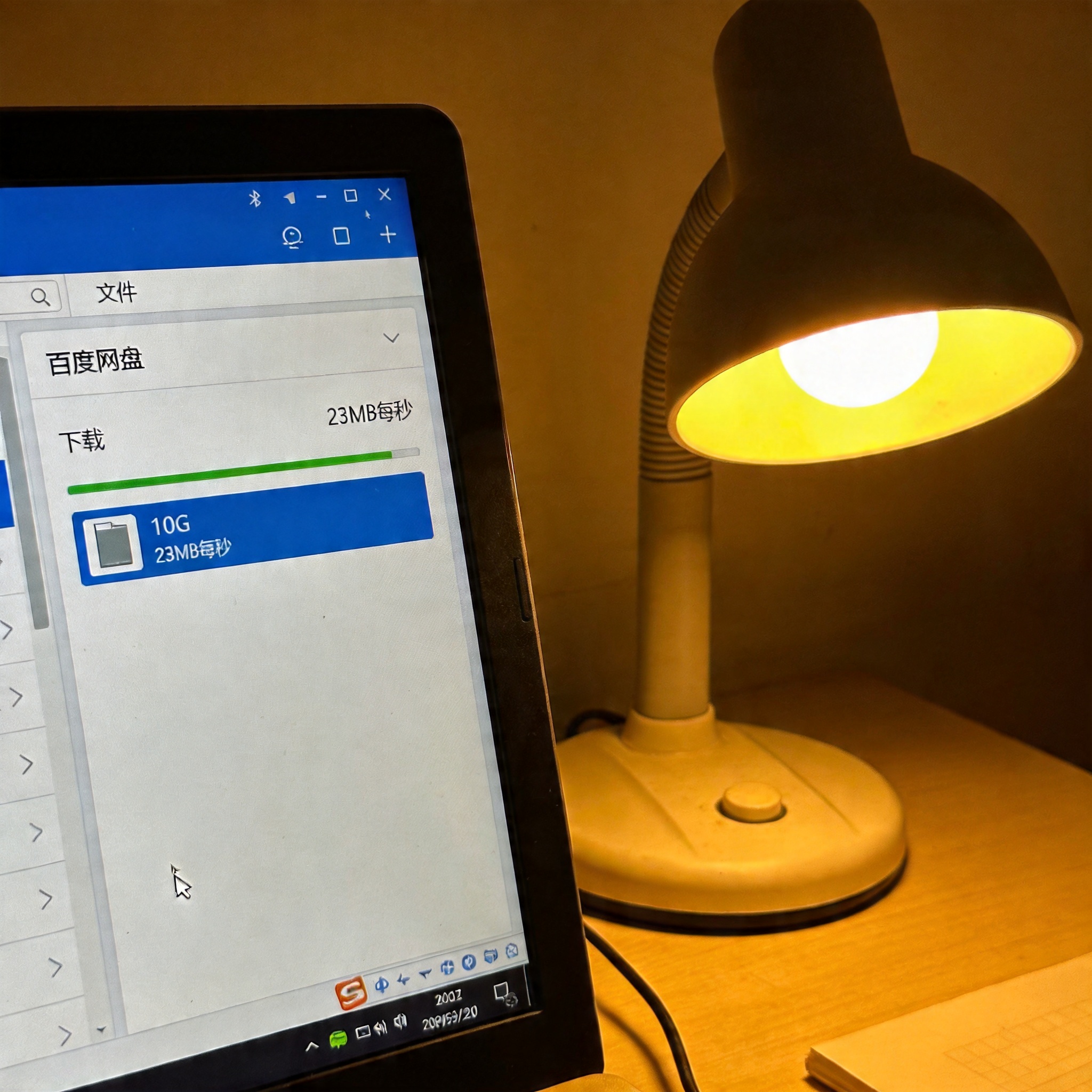 电脑屏幕显示百度网盘文件列表，其中10G大小的文件正在下载，速度显示23MB每秒，旁边摆着桌面台灯，暖光