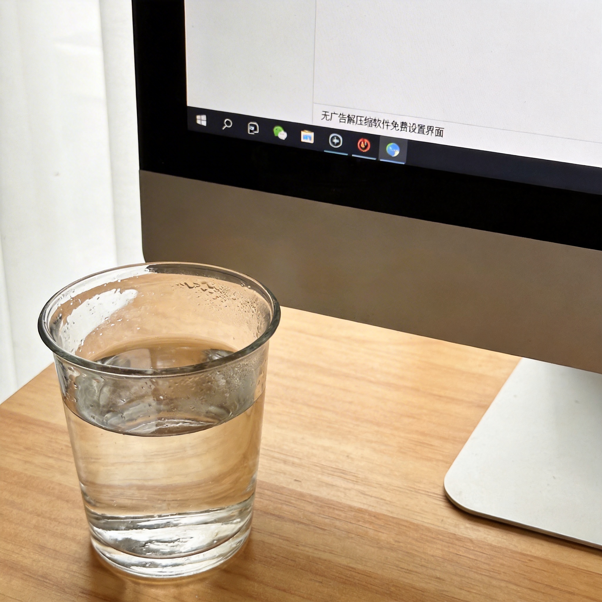 书桌上的主机，屏幕显示无广告解压缩软件免费设置界面，桌角放着水杯，自然光