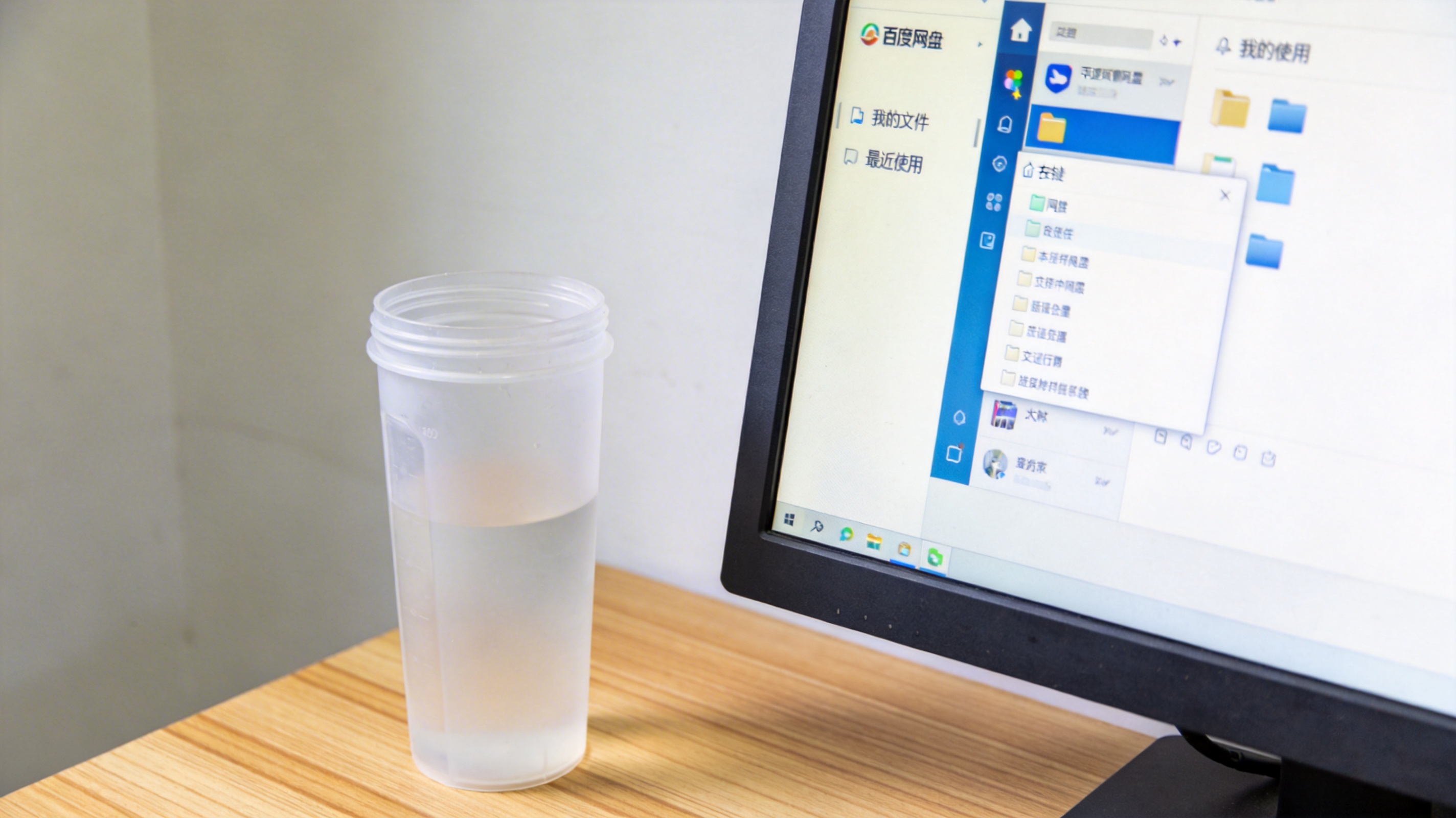 电脑桌的台式电脑，屏幕显示百度网盘客户端界面，右键菜单展开在文件上方，旁边放着塑料水杯，室内自然光