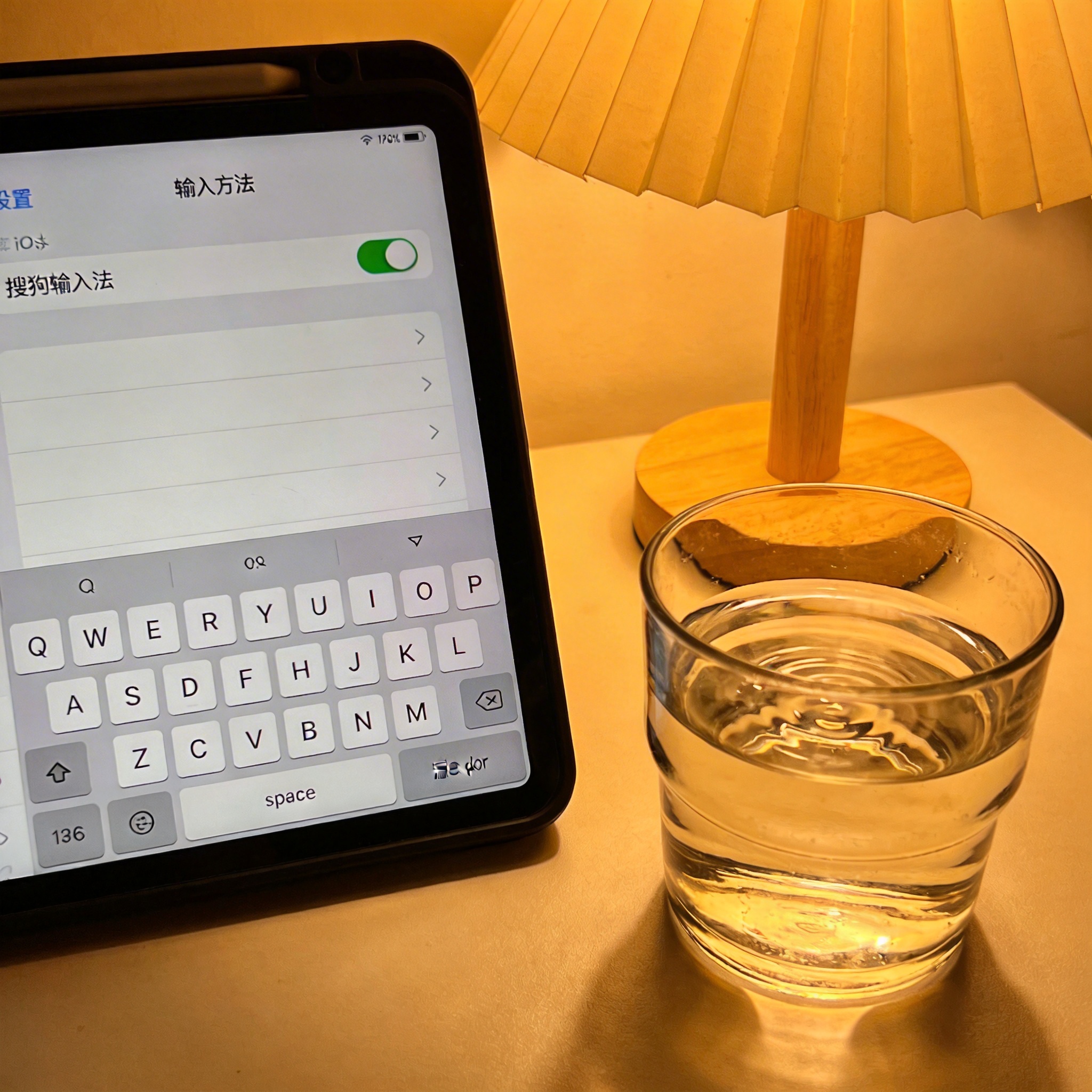桌面上的iPad，屏幕显示iOS系统的输入法设置界面，已选中搜狗输入法，旁边放着半杯温水，台灯暖光