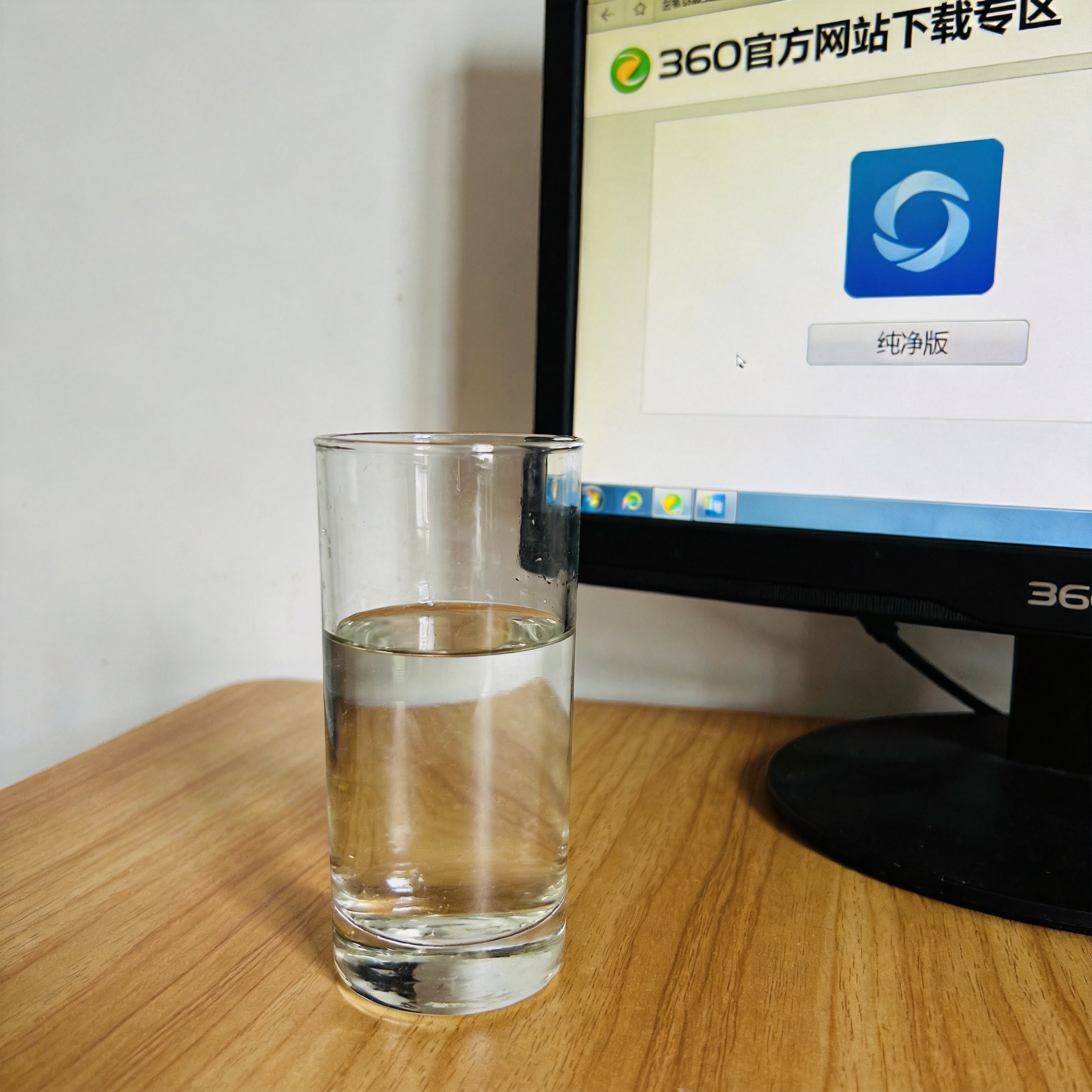 电脑桌的电脑屏幕显示360官方网站下载专区，页面标注着“纯净版”选项，旁边放着半杯凉白开，室内自然光