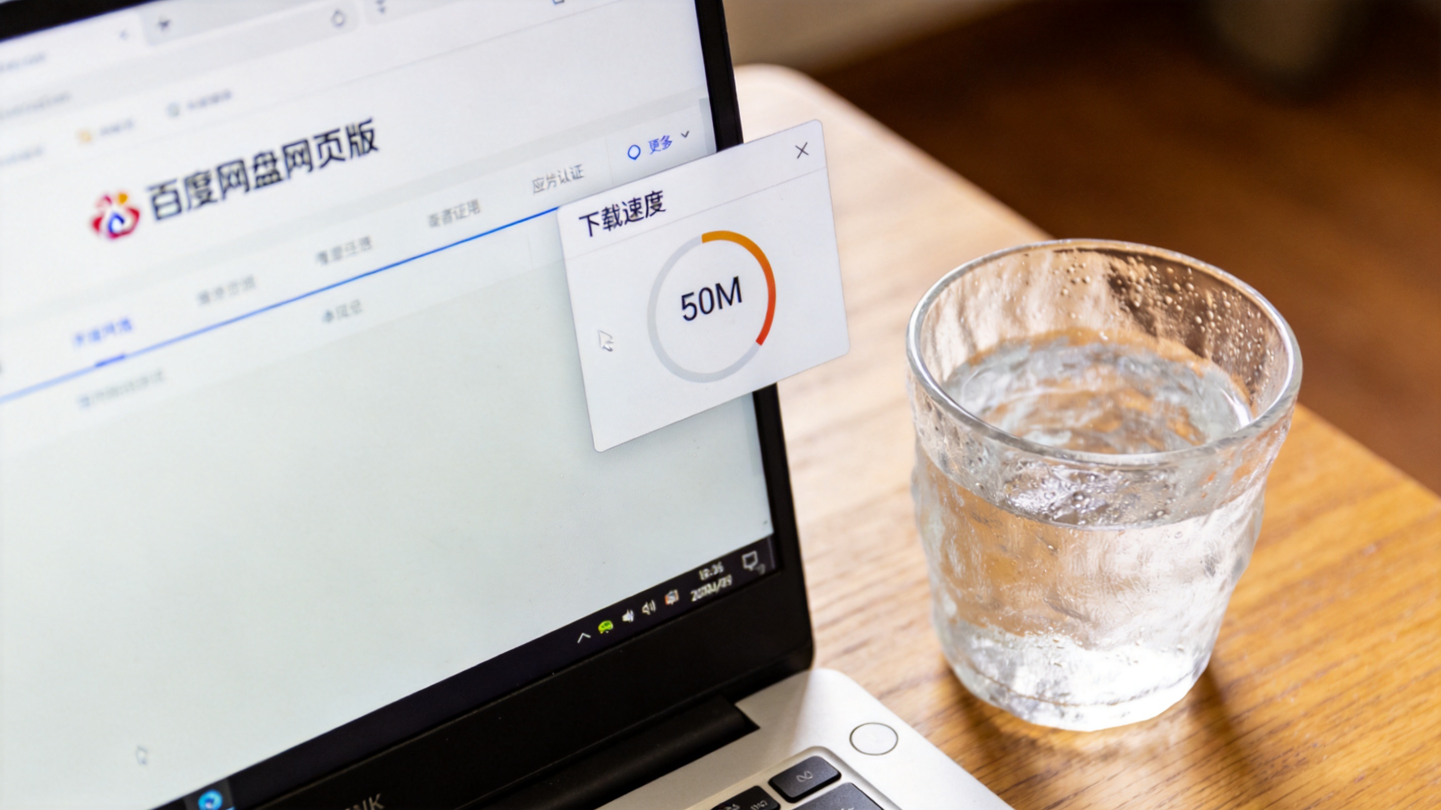 电脑桌的电脑屏幕显示百度网盘网页版界面，右侧悬浮窗显示下载速度50M，旁边放着半杯凉水，室内自然光