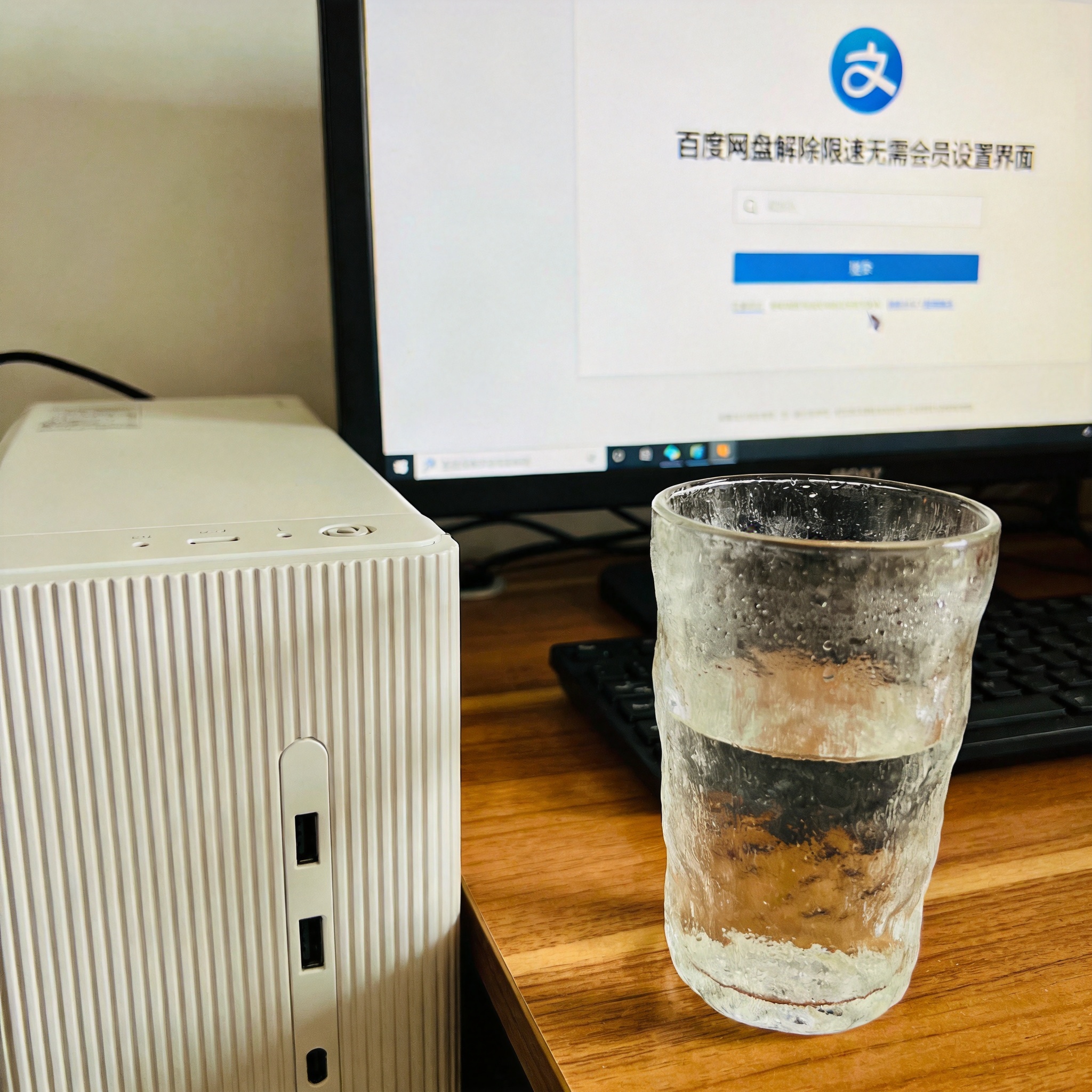 书桌上的主机，屏幕显示百度网盘解除限速无需会员设置界面，桌角放着水杯，自然光
