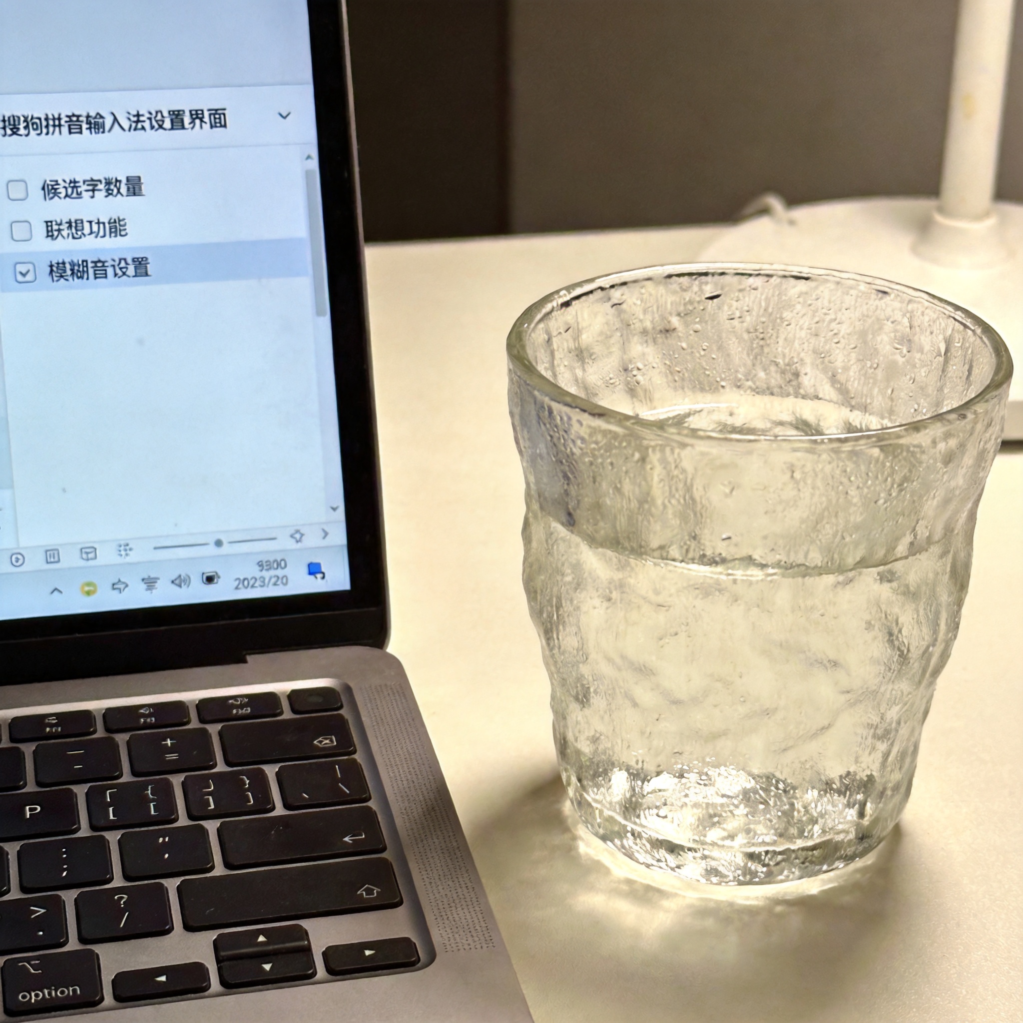 电脑屏幕显示搜狗拼音输入法设置界面，页面上有候选字数量、联想功能、模糊音设置的选项，旁边放着半杯温水