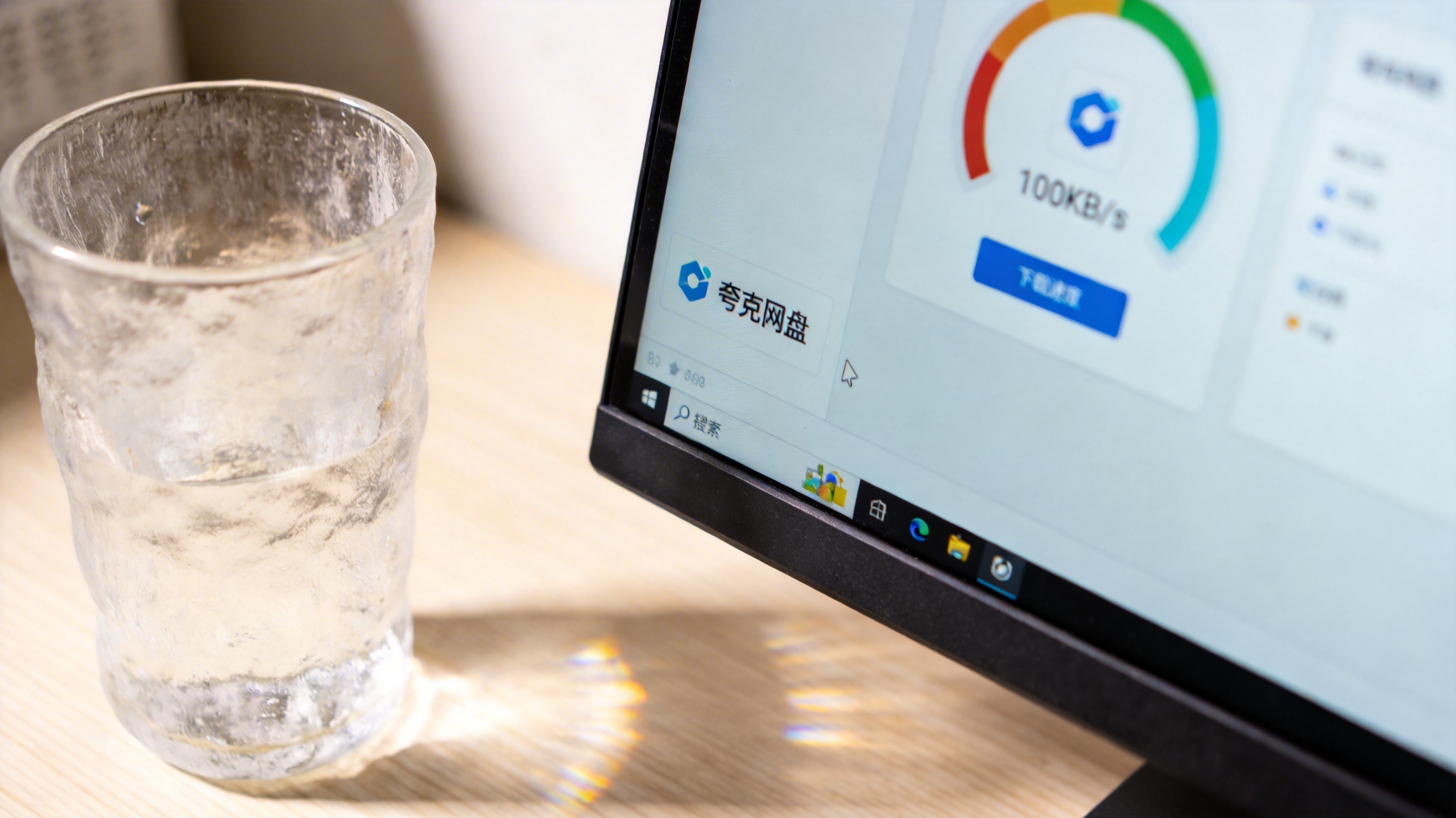 电脑桌面，屏幕显示夸克网盘客户端界面，下载速度显示每秒100KB，旁边放着半透明水杯，室内自然光