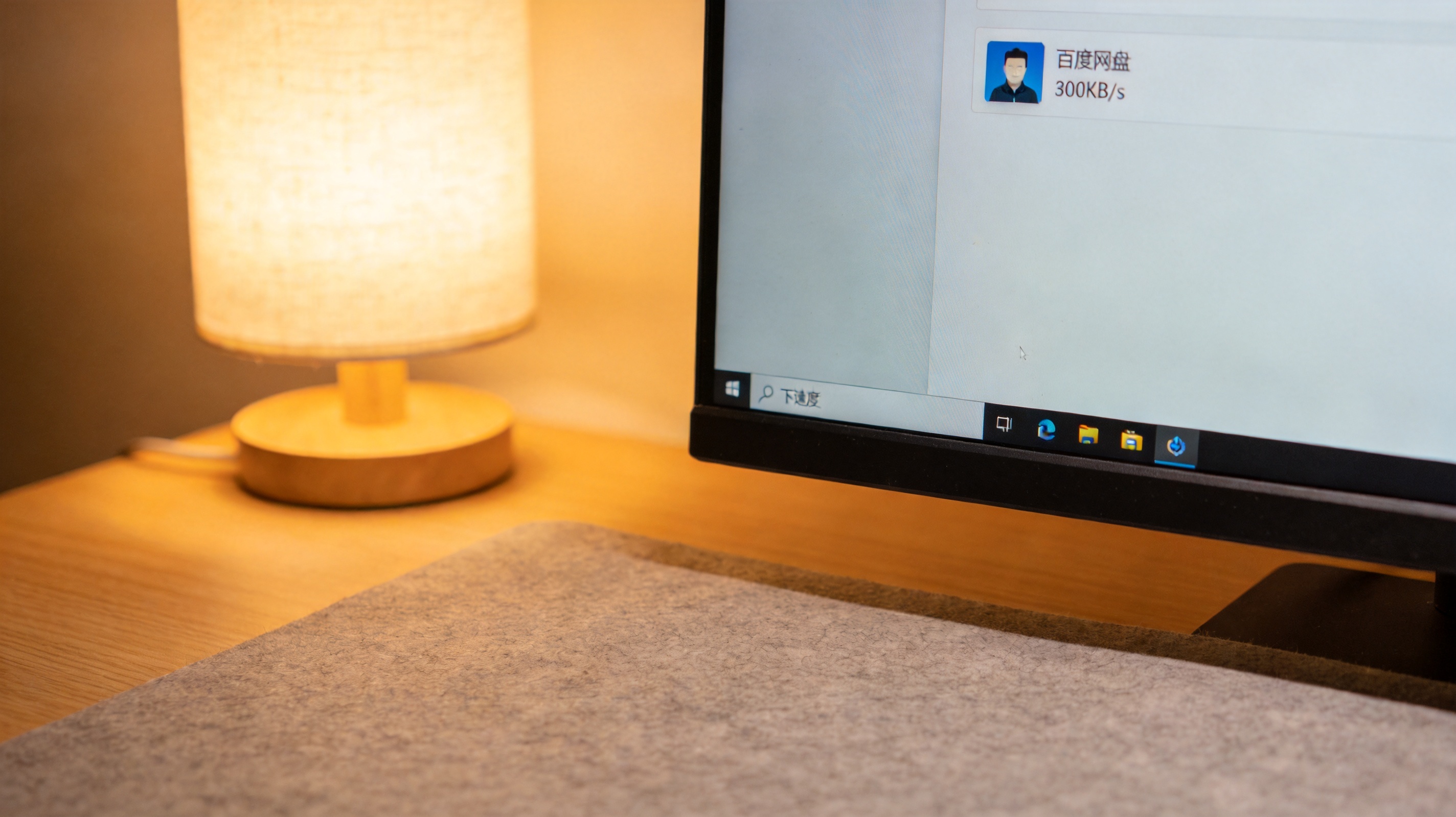 电脑屏幕显示百度网盘下载任务界面，单个任务下载速度显示300KB/s，桌面铺着浅灰色桌垫，台灯暖光