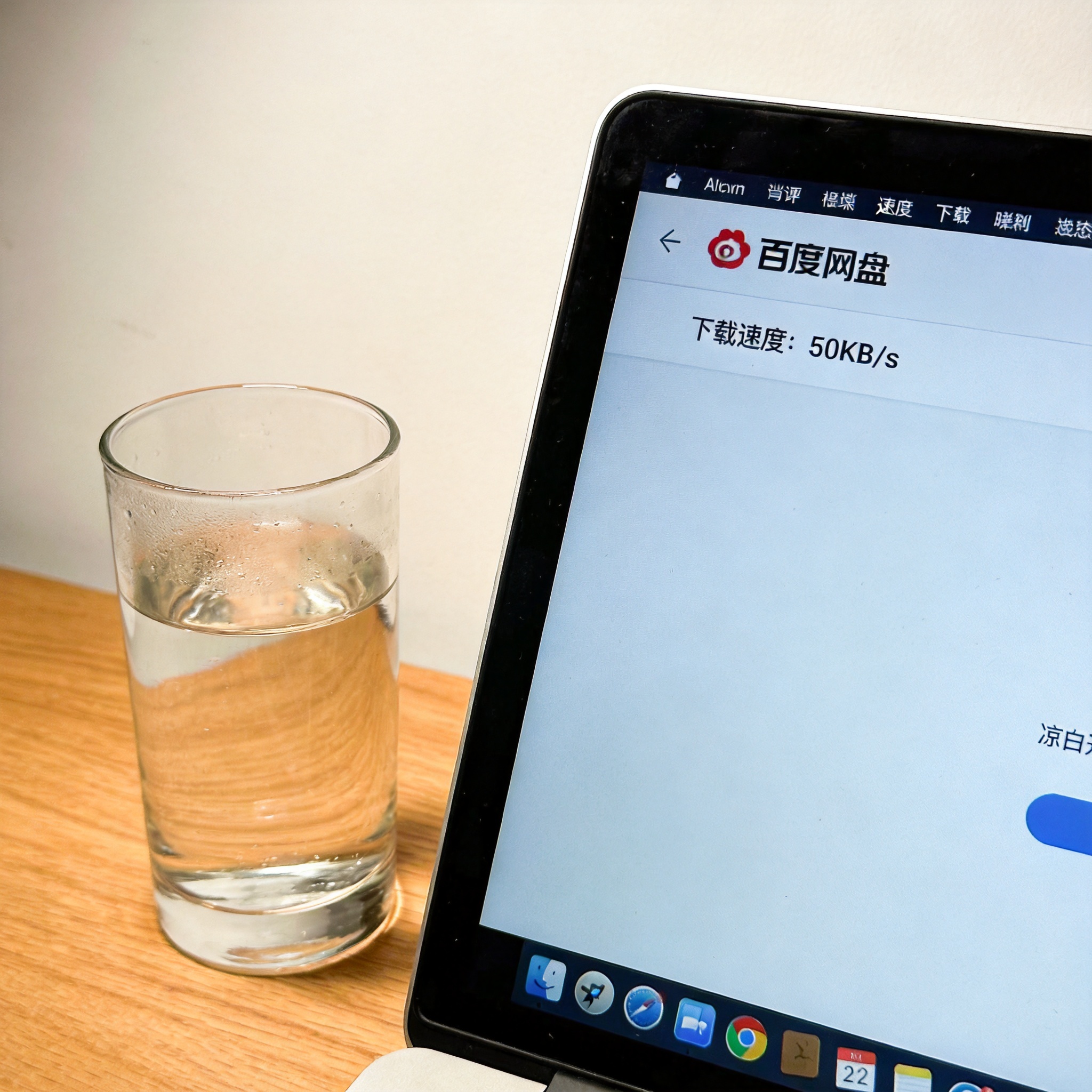 电脑桌的电脑屏幕显示百度网盘下载界面，下载速度显示50KB/s，旁边放着半杯凉白开，室内自然光