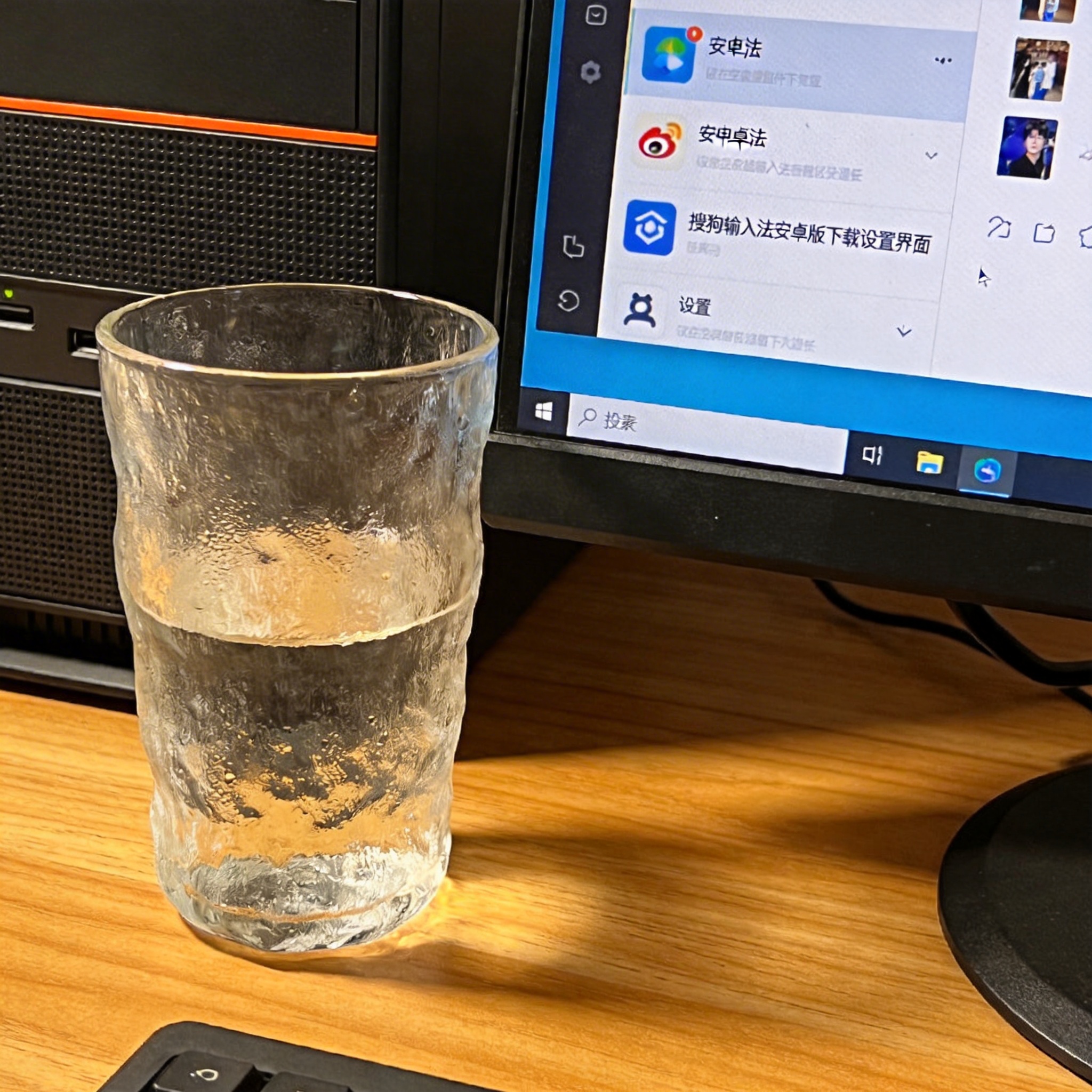 书桌上的主机，屏幕显示搜狗输入法安卓版下载设置界面，桌角放着水杯，自然光