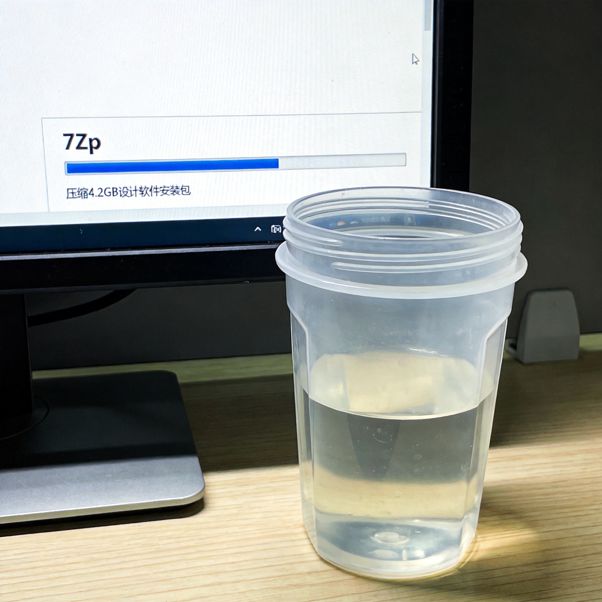 台式机屏幕显示7Zip压缩进度条，正在压缩4.2GB的设计软件安装包，旁边放着半透明塑料水杯，屏幕光