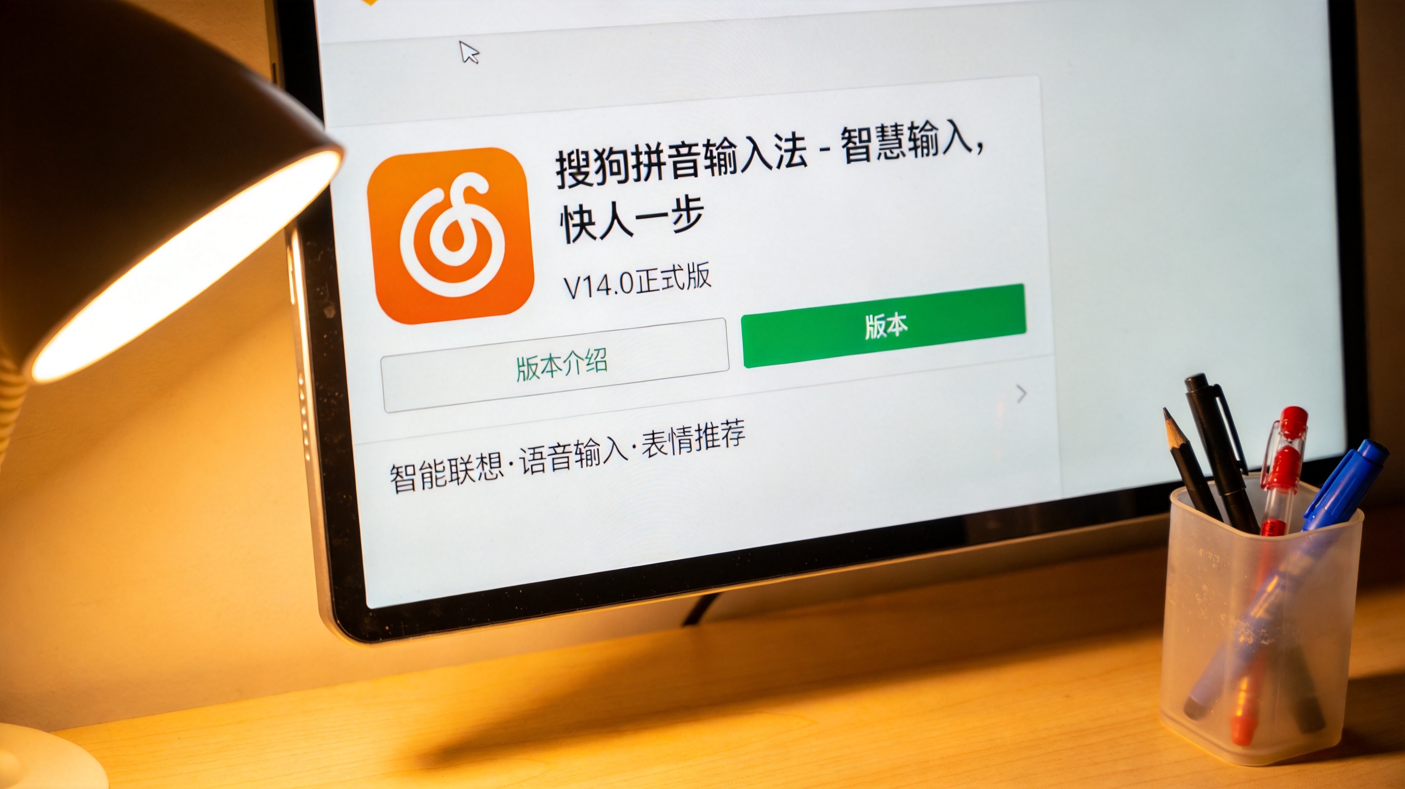 台式机桌面，屏幕显示搜狗拼音输入法官网的版本介绍页面，桌面角落有个塑料笔筒，台灯暖光