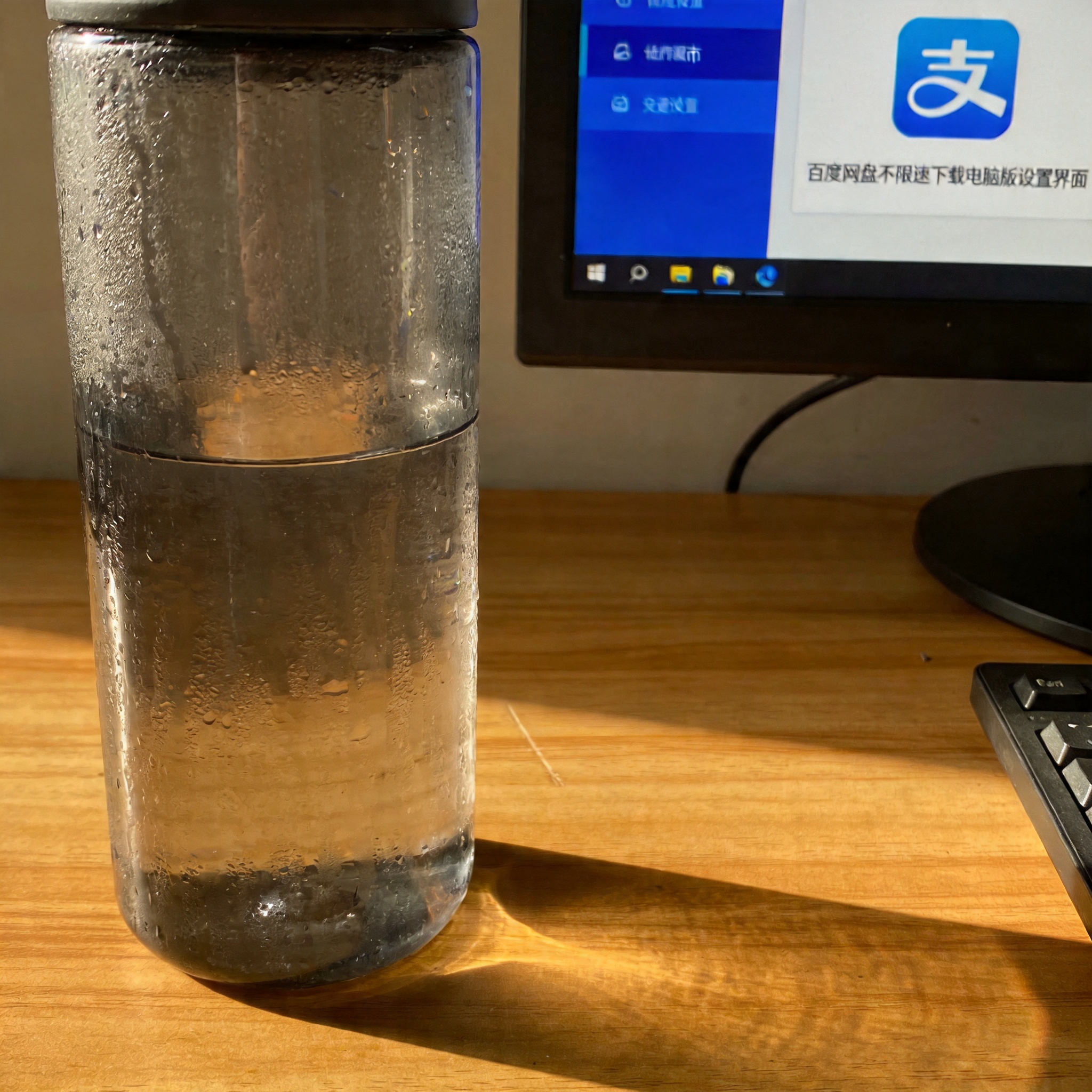 书桌上的主机，屏幕显示百度网盘不限速下载电脑版设置界面，桌角放着水杯，自然光