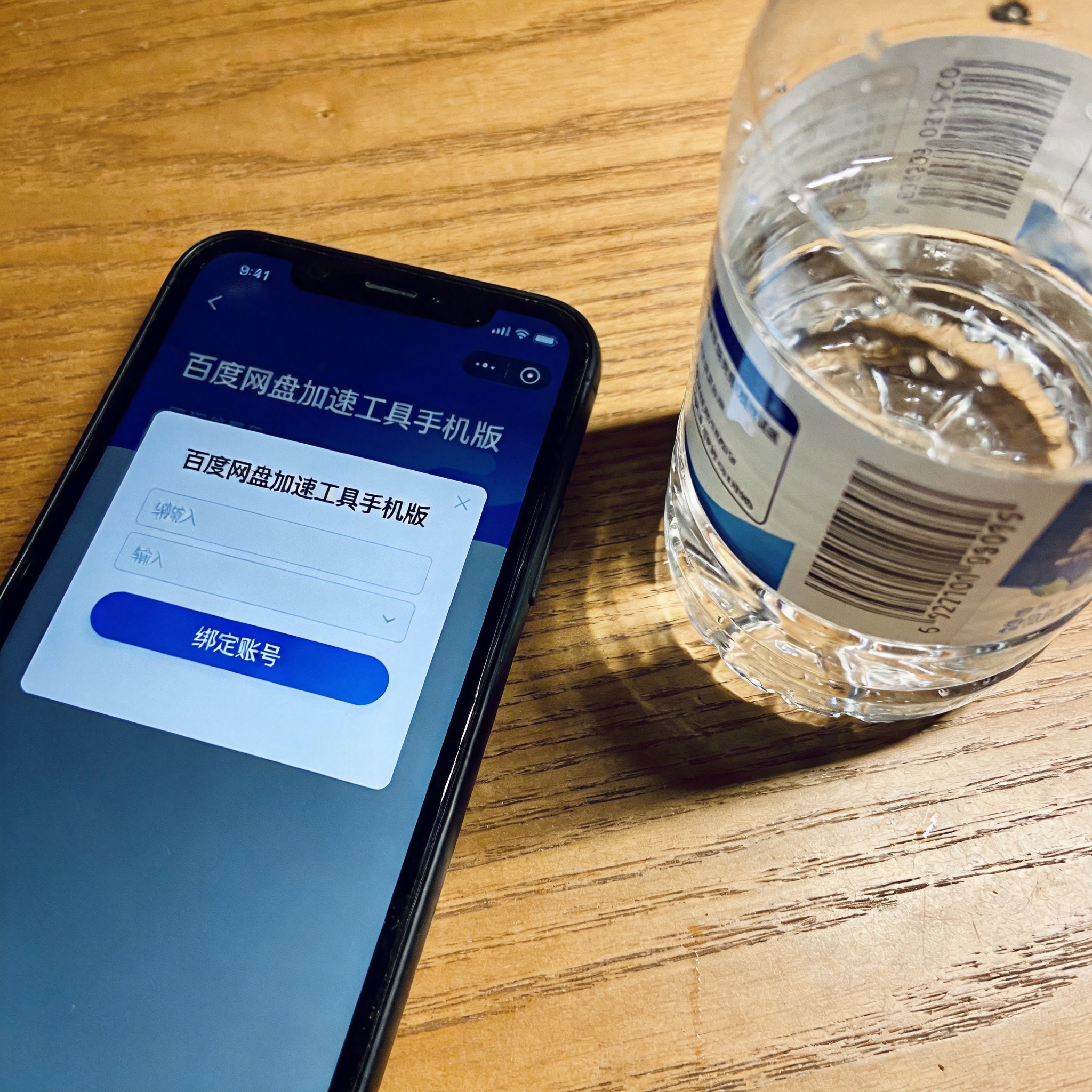 桌面放着的手机，屏幕显示百度网盘加速工具手机版的绑定账号窗口，旁边有半瓶矿泉水，室内自然光