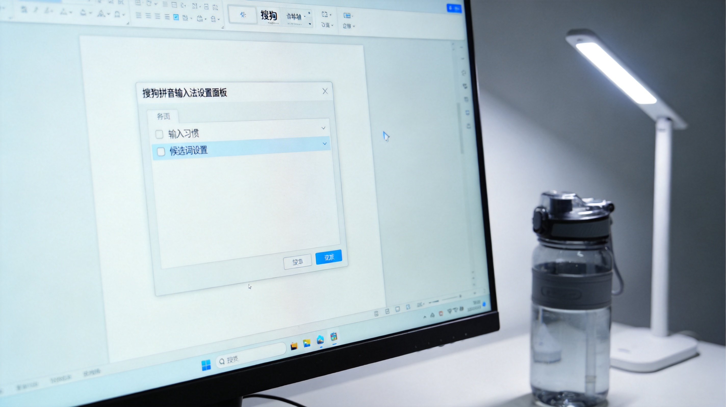 电脑屏幕显示搜狗拼音输入法设置面板，面板里有输入习惯、候选词设置选项，旁边放着塑料水杯，台灯冷光